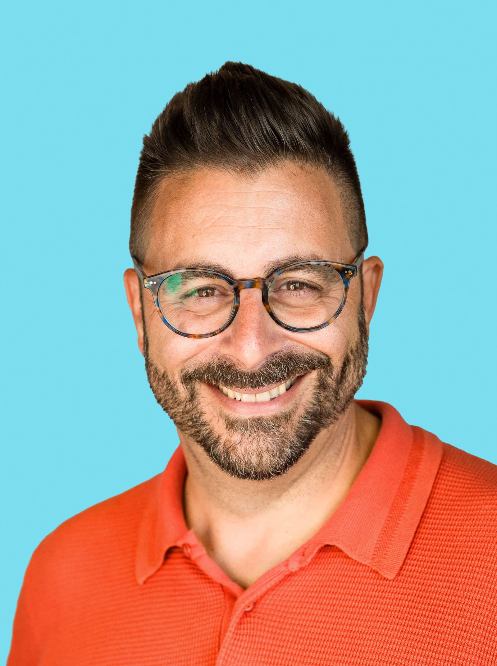 A man with short dark hair, a beard, and glasses smiles at the camera. He is wearing a bright orange polo shirt and stands against a solid light blue background.