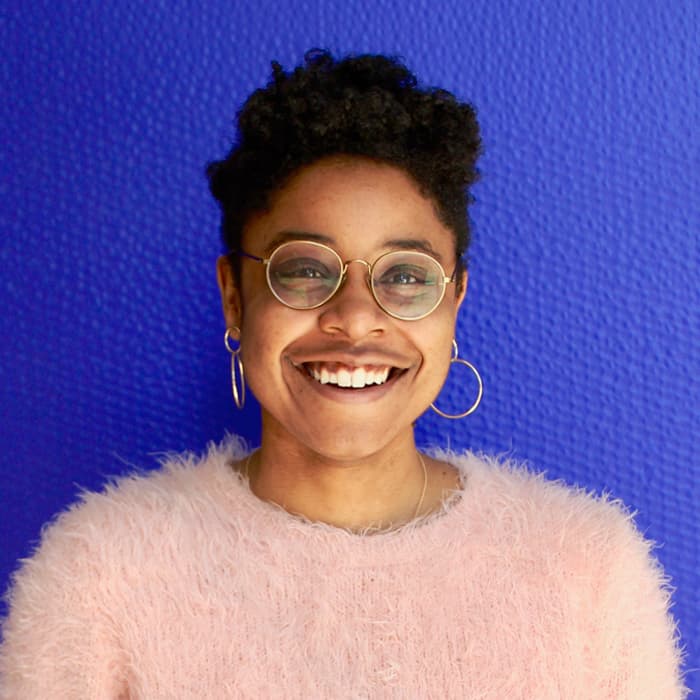 A person with short curly hair, wearing round glasses and hoop earrings, smiles warmly at the camera. They are dressed in a fuzzy light pink sweater and stand against a vibrant blue textured background.