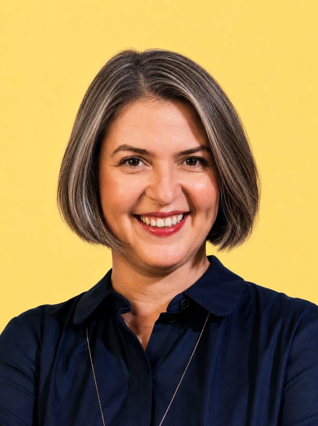 A woman with short brown and gray hair smiles, wearing a dark blue collared shirt and a necklace, standing in front of a bright yellow background.