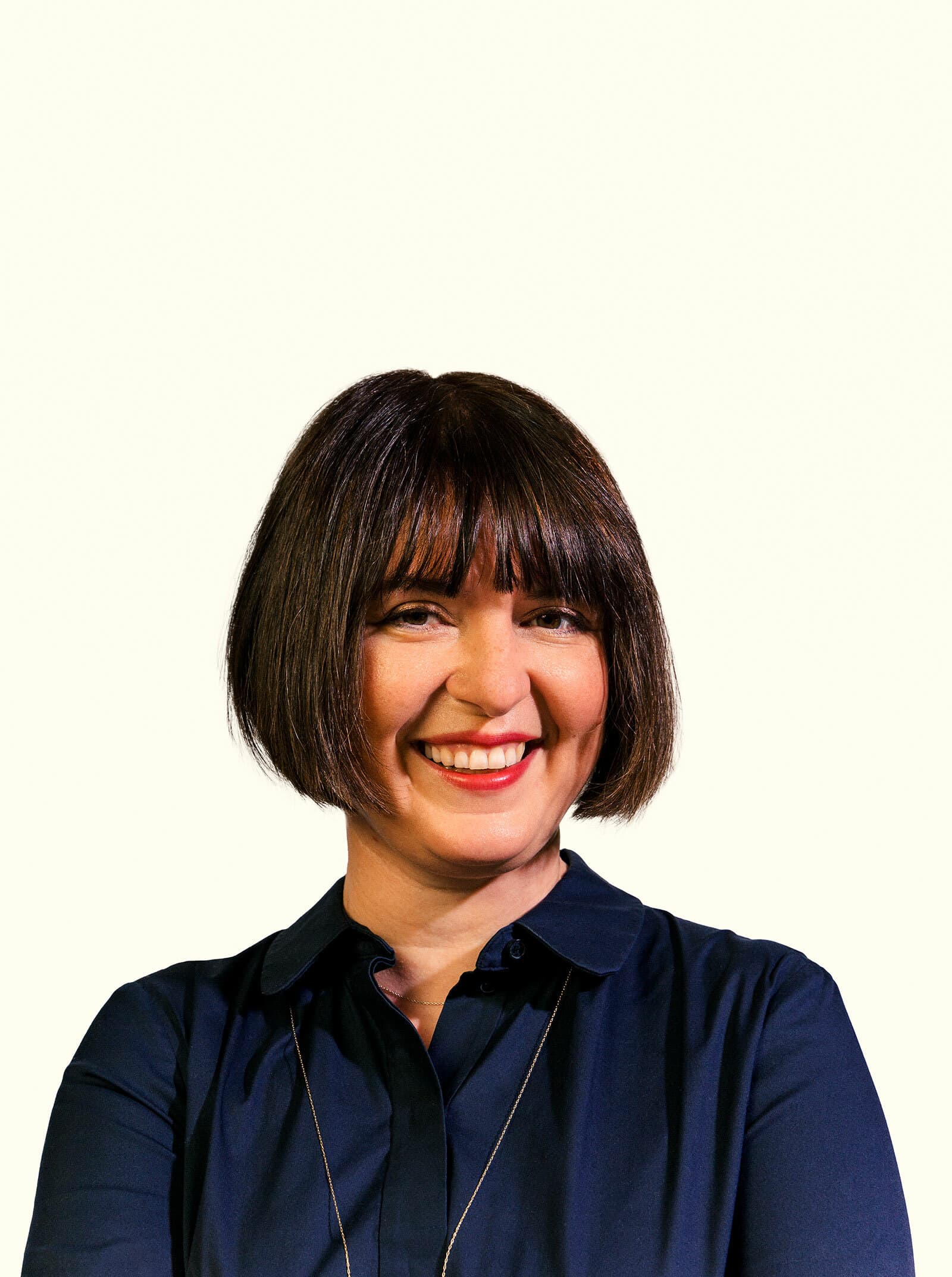 A woman with a short bob haircut and bangs is smiling at the camera. She is wearing a dark navy collared shirt, and the background is a plain, light color.
