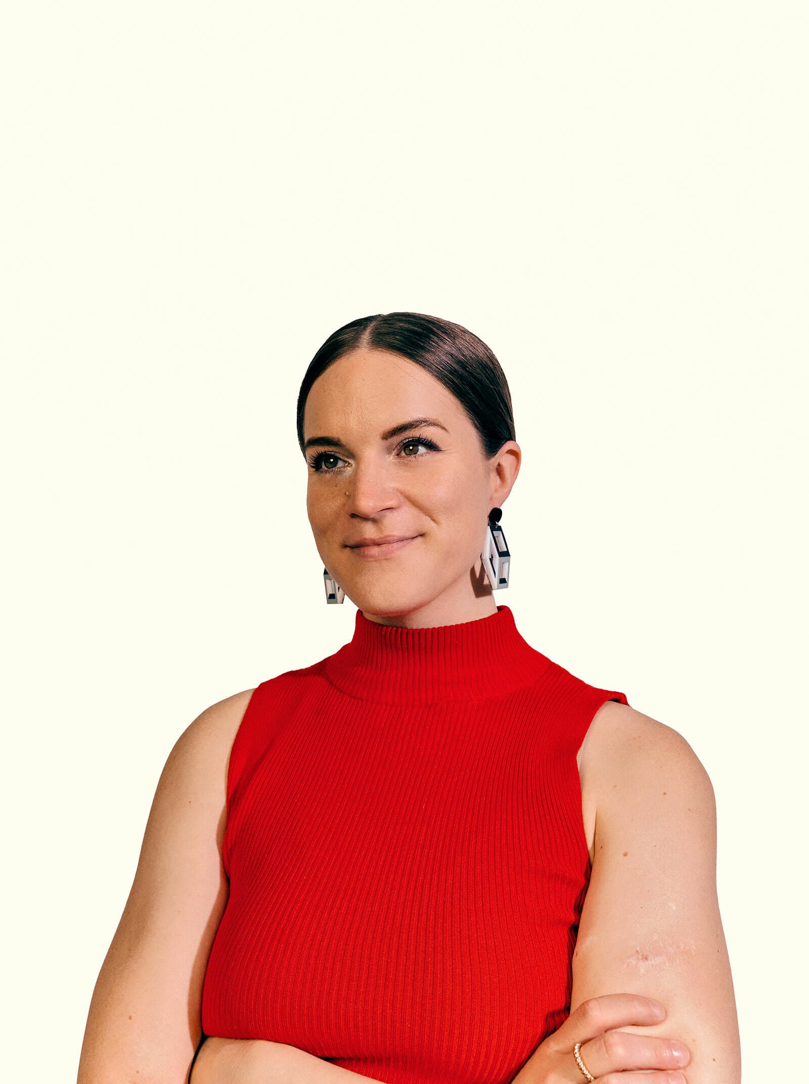 A person with straight dark hair, neatly parted in the middle, wearing geometric earrings and a sleeveless, high-neck red top. They are looking slightly upwards with a closed-mouth smile and have their arms crossed. The background is plain light beige.