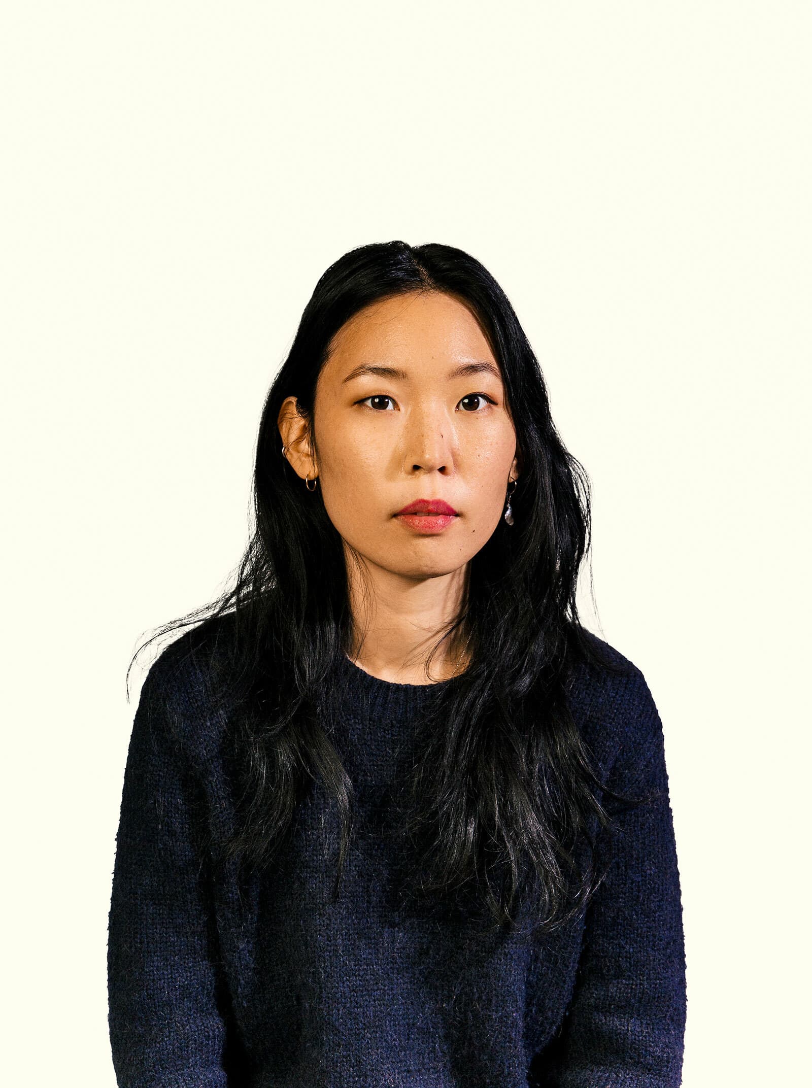 A person with long, black hair and wearing a dark sweater is looking directly at the camera against a plain, light-colored background. The individual has a neutral expression with a calm demeanor.