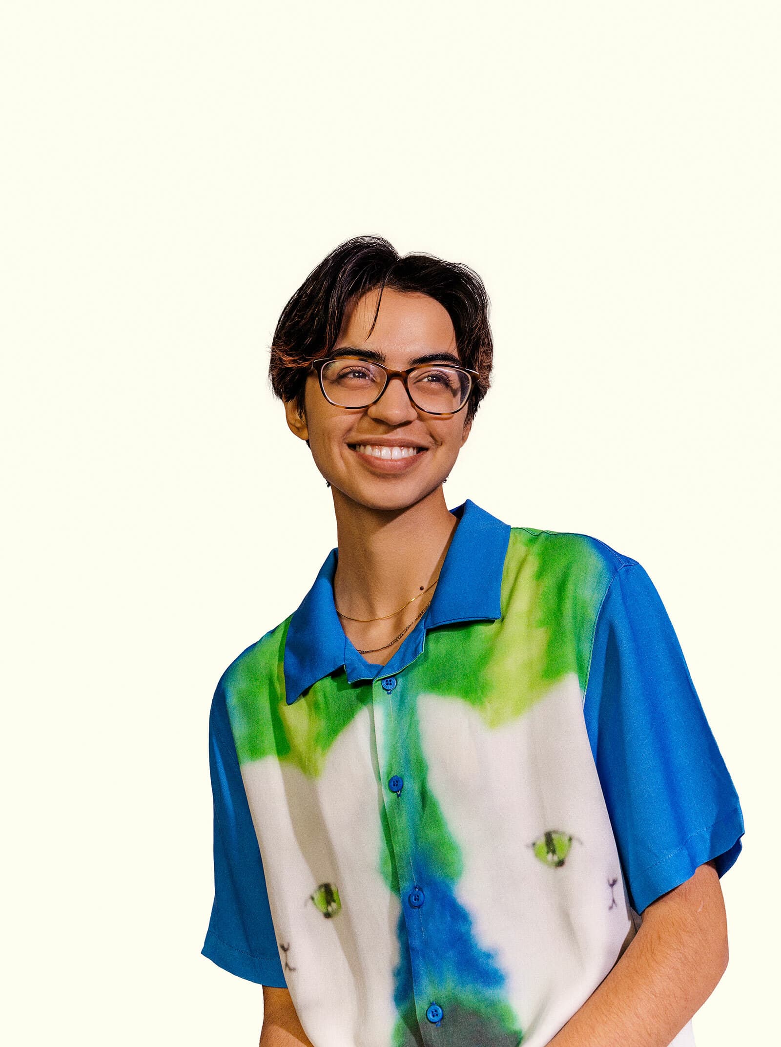 A person with short hair, wearing glasses and a colorful shirt with green, blue, and white patterns smiles while looking off to the side. The background is plain and light-colored.