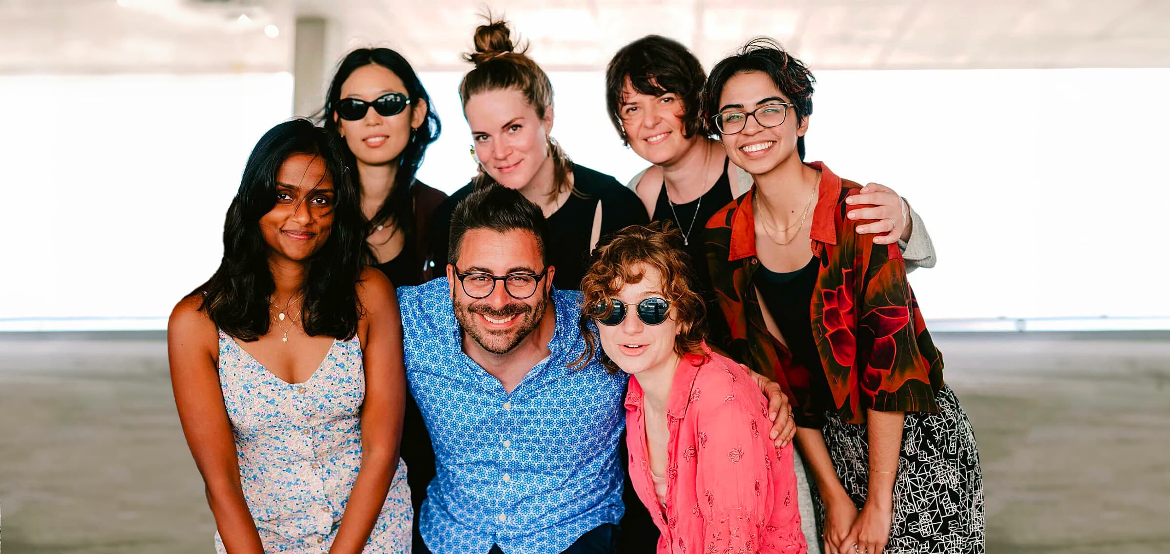 A group of seven smiling people are huddled together for a photo. They are indoors, and natural light fills the background. The group includes four women and three men, all casually dressed and appearing relaxed and happy.