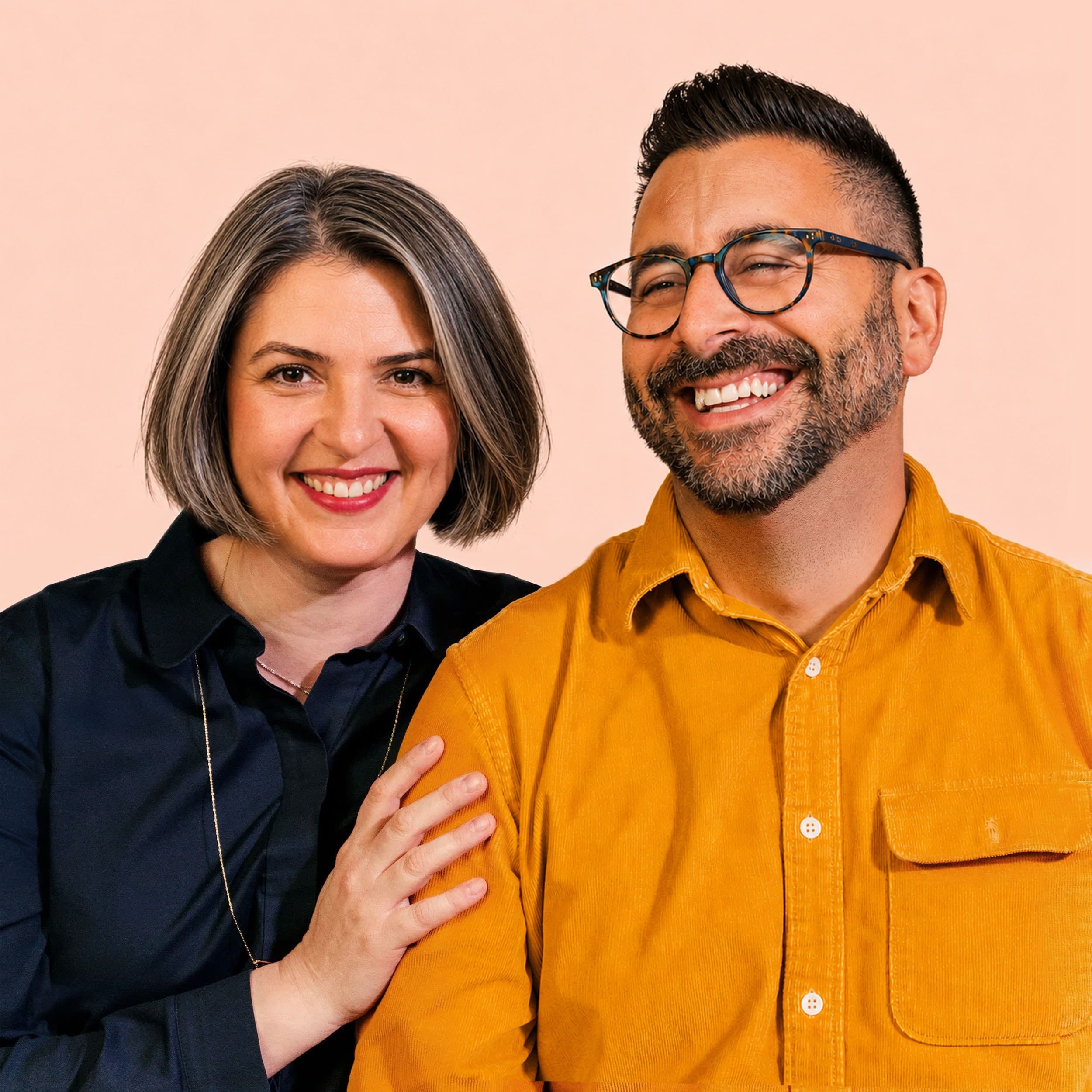 Two people smiling, one with short brown hair wearing a black shirt, the other with short dark hair, glasses, and a mustard yellow shirt. They are standing close together against a light peach background.