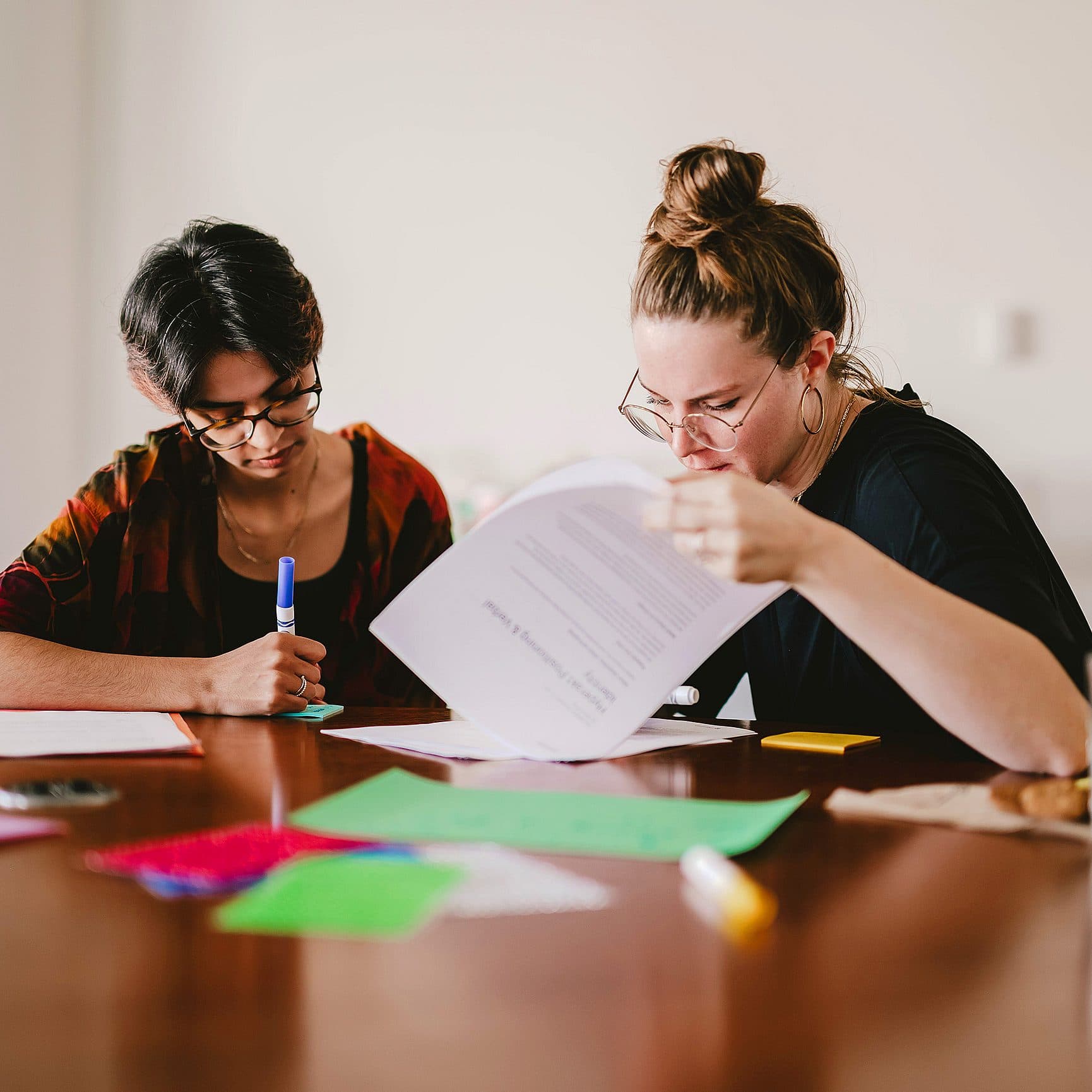 Two individuals sit at a wooden table working together. One person holds a pen and the other reviews a document. Brightly colored papers are scattered on the table. Both look focused and engaged. Glasses and a cup are also visible on the table.