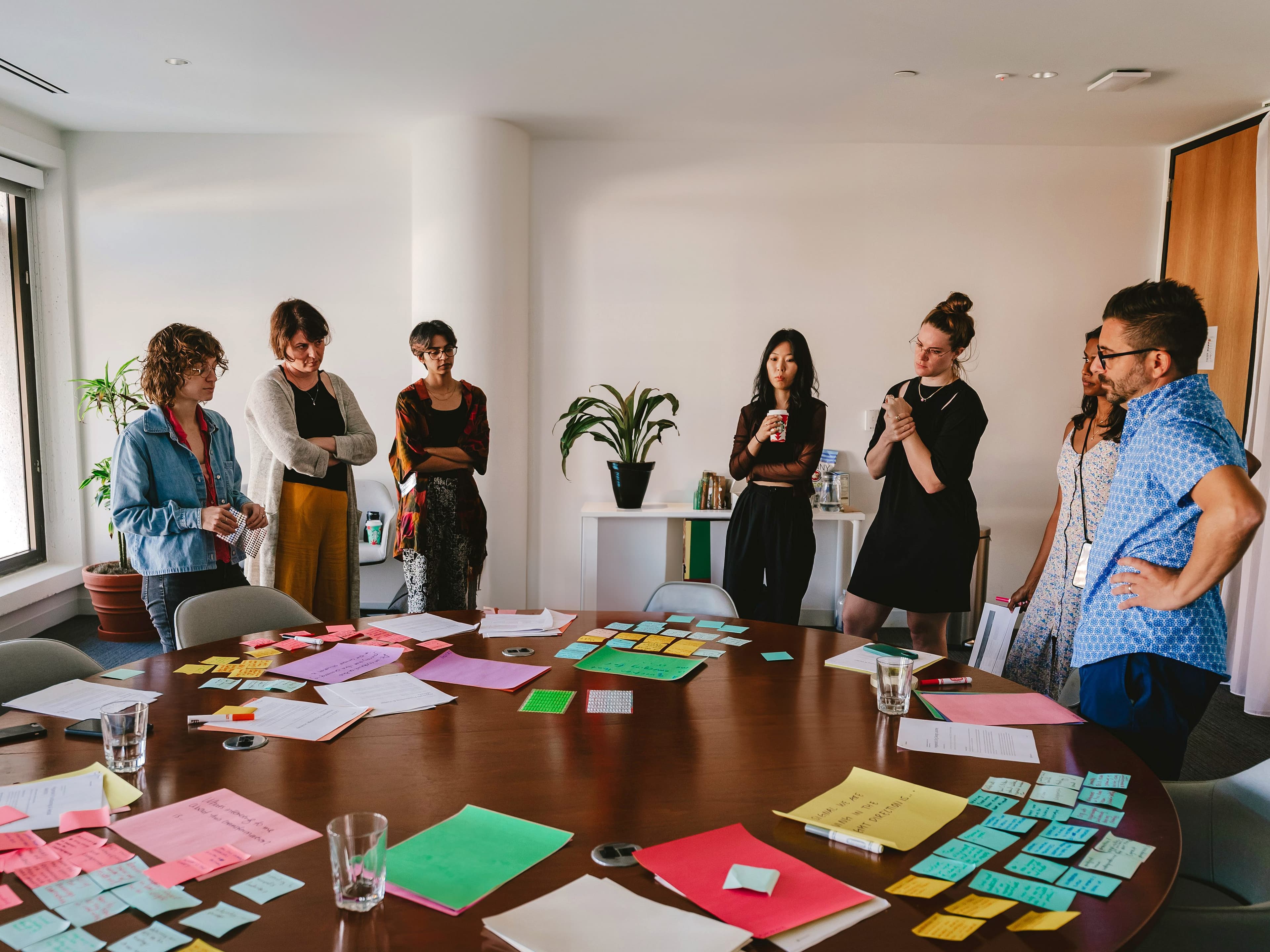 A group of seven people are gathered around a round table covered with colorful sticky notes, papers, and glasses of water in a brightly lit office. Some are standing, while others are engaged in discussion, suggesting a brainstorming session or meeting.