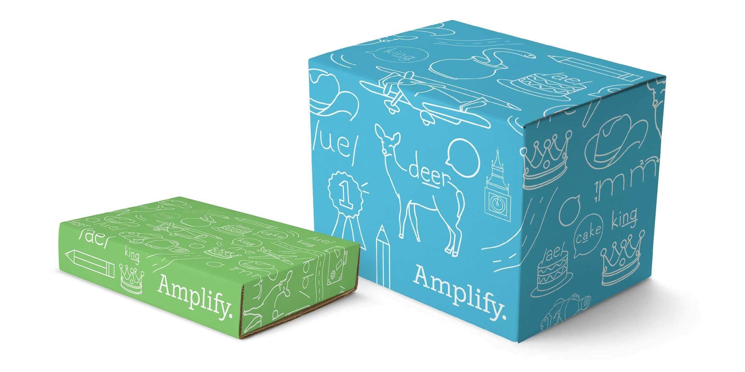 Two cardboard boxes with illustrated designs stand side by side. The larger box is blue, covered in white doodles depicting various objects and labeled with "Amplify." The smaller box is green, with similar white doodles and the same "Amplify" text.