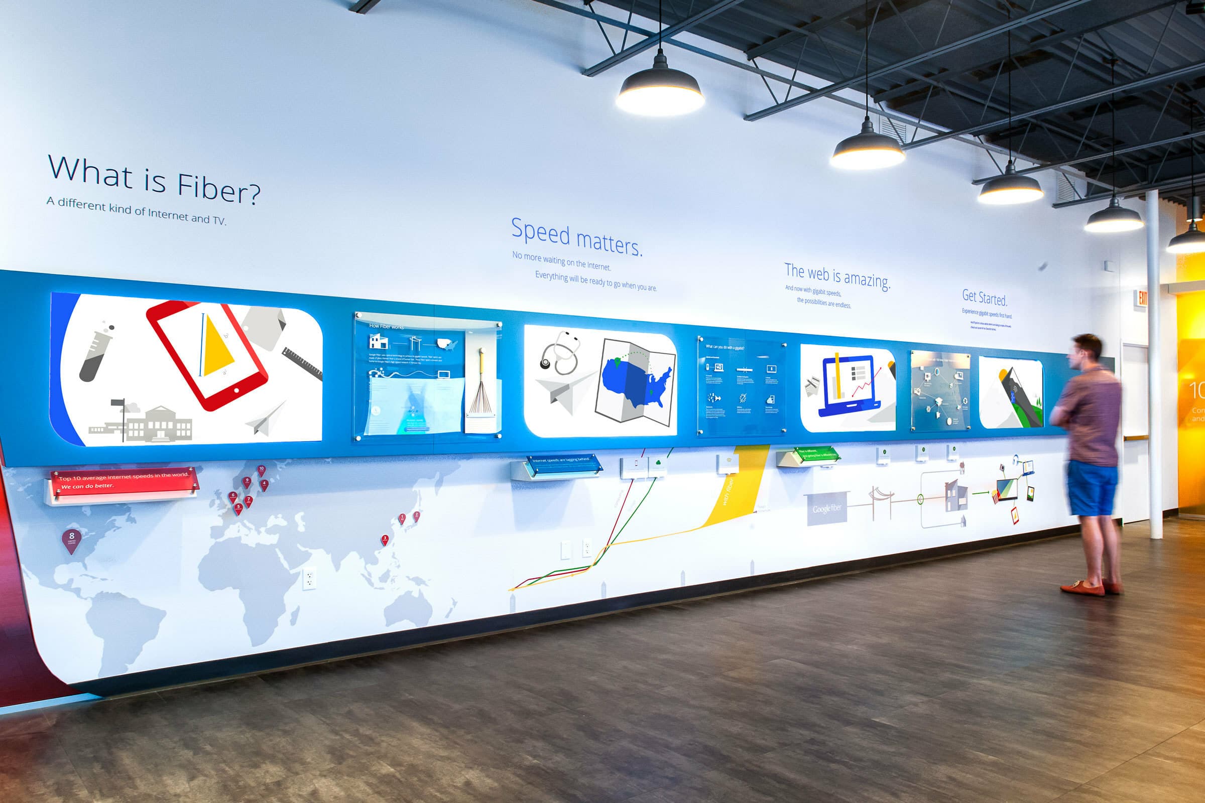 A person stands in front of an interactive display wall explaining fiber internet technology. The wall features visual aids, maps, text, and interactive screens under categories such as "What is Fiber?", "Speed matters," "The web is amazing," and "Get Started.