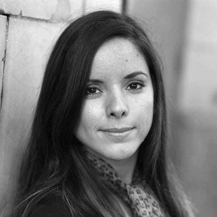 A black and white close-up photo of a woman with long, straight hair. She is looking directly at the camera with a neutral expression. She is wearing a patterned scarf and a dark top. The background is slightly blurred, suggesting an outdoor setting.
