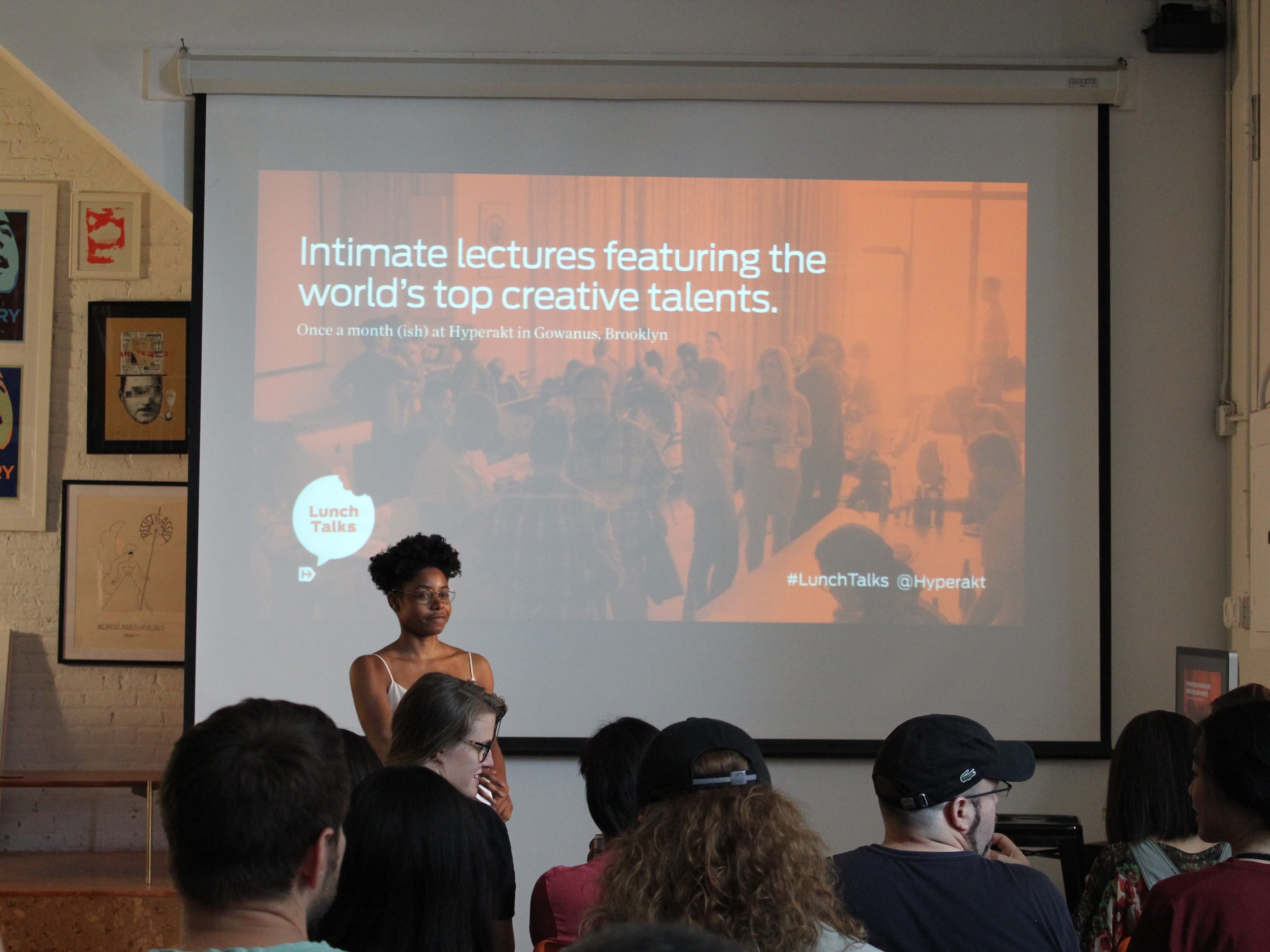 A woman stands in front of a crowd in a room, speaking at an event. Behind her, a screen displays a presentation titled "Intimate lectures featuring the world's top creative talents." The audience is seated and attentively watching the speaker.