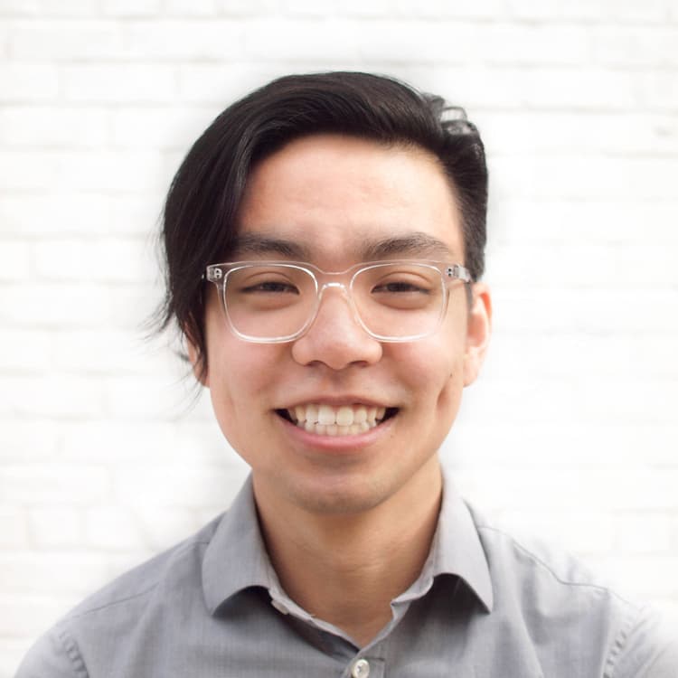A young man with clear-framed glasses and dark, side-swept hair smiles warmly at the camera. He is wearing a light gray button-up shirt and stands in front of a white brick background.