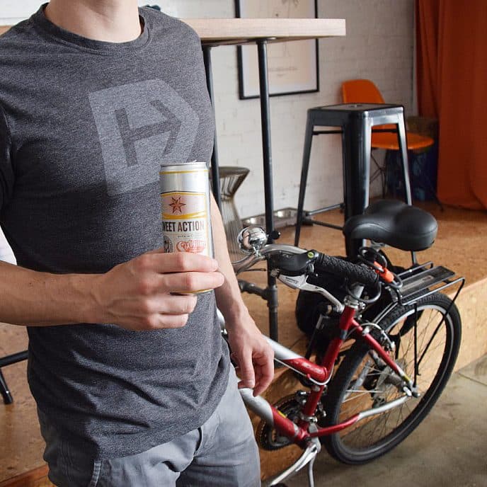 A person in a grey t-shirt holding a can of "Sweet Action" drink stands next to a red bicycle indoors. The background shows a bar-like setting with stools, a counter, and an orange curtain.