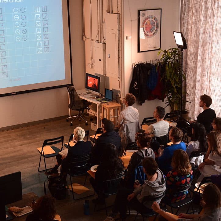 A person stands and points to a projected presentation on a screen. An audience is seated and attentively watching. The room has light wooden floors, a desk with computers, some wall art, and curtains drawn to the right side.