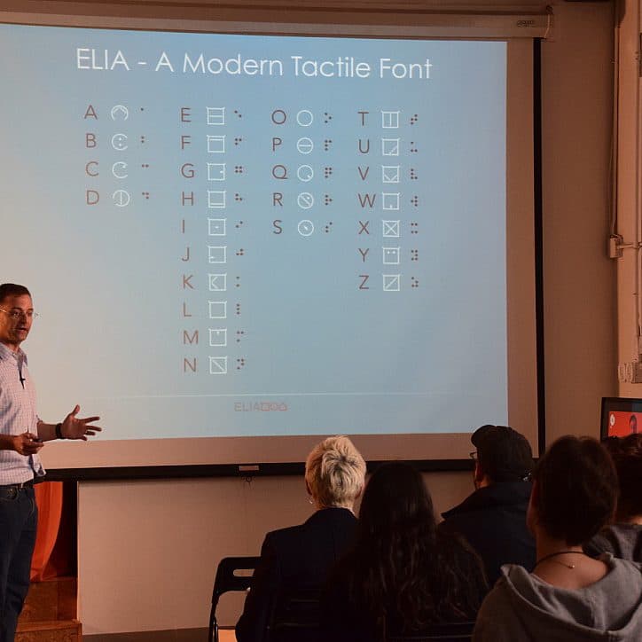 A man in a blue shirt stands at the front of a room, speaking to an audience. Behind him is a projected slide titled "ELIA - A Modern Tactile Font," showing a chart of tactile symbols. The room is dimly lit, with several audience members visible from the back.