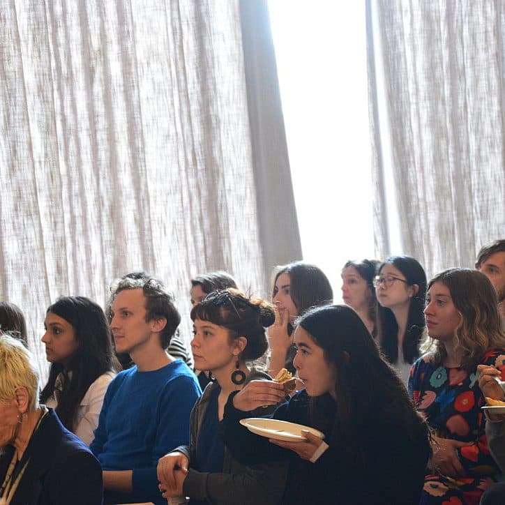 A group of people seated closely together, attentively listening. They are in a room with large curtains letting in light. Some individuals are eating or holding plates. The audience displays a mix of casual and focused expressions.