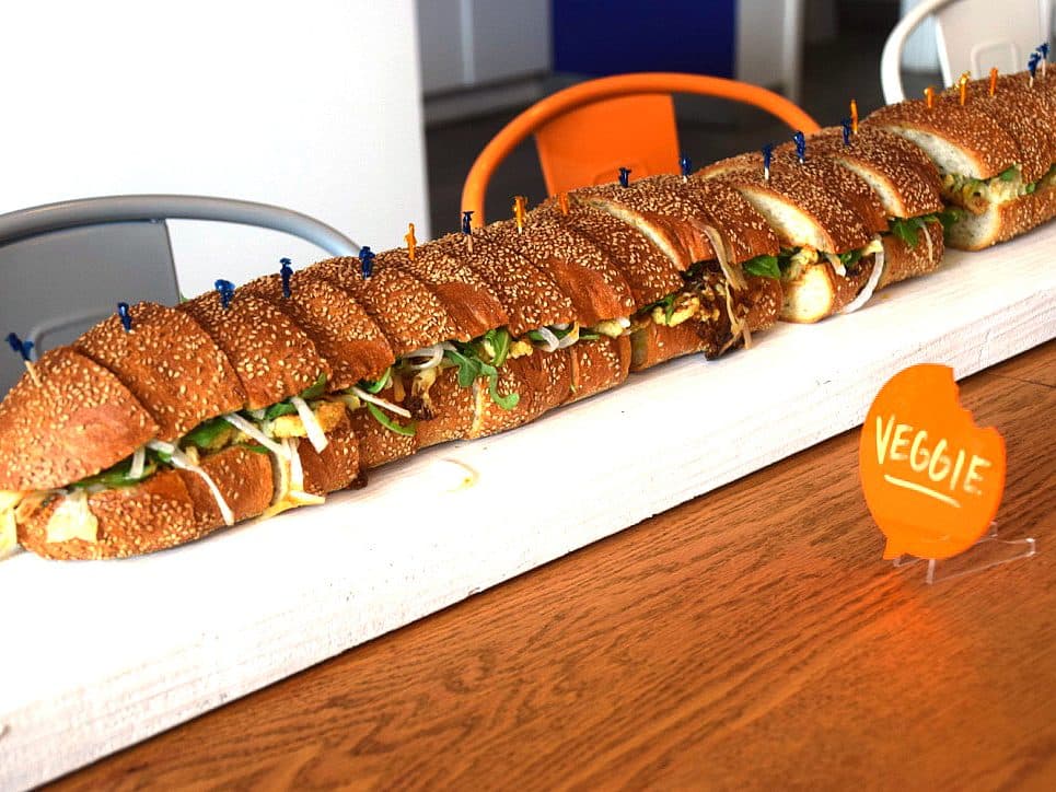 A long sandwich cut into multiple sections is placed on a wooden board. It is labeled "VEGGIE" with a small orange sign and is filled with fresh vegetables. The sandwich is secured with colorful toothpicks, and there are chairs and a table in the background.