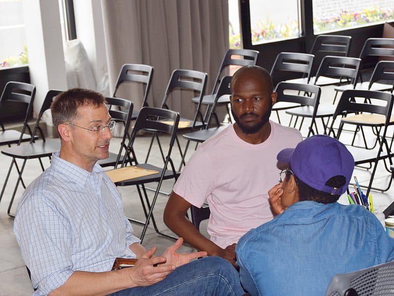 Three people sit in conversation in a room with rows of empty black chairs. One person is gesturing with hands, suggesting they are speaking, while the other two listen attentively. The room has large windows and curtains.