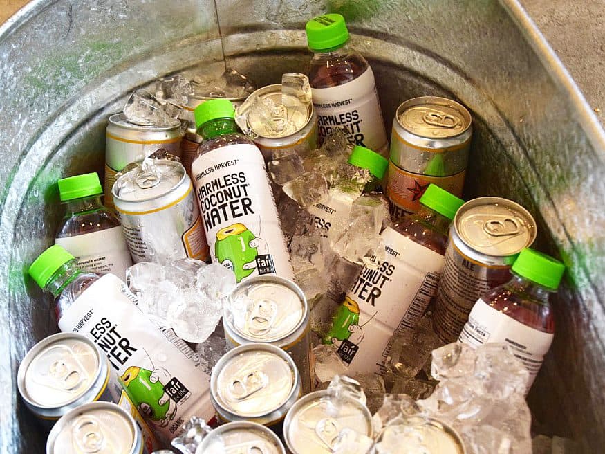 A metal tub filled with ice, containing bottles of Harmless Harvest Coconut Water and cans of Spindrift Sparkling Water. The drinks are chilled and arranged chaotically within the ice.
