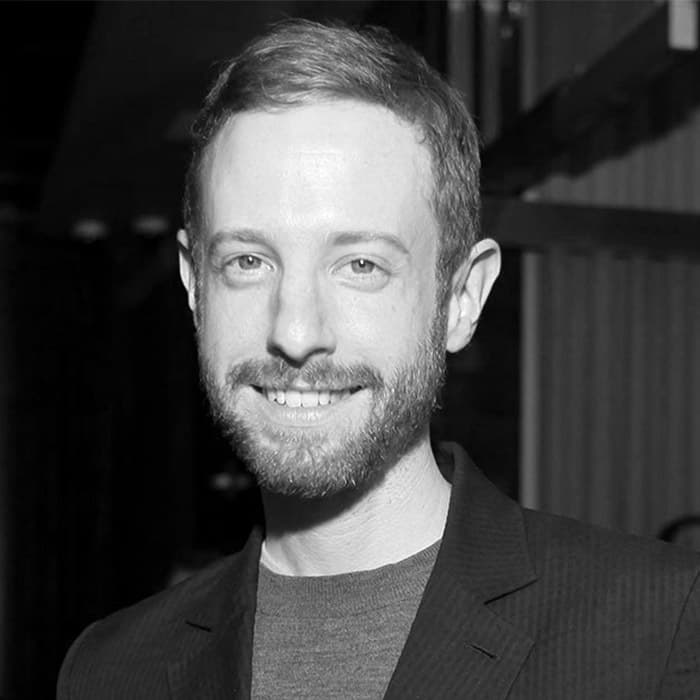 Black and white photo of a smiling man with short hair and a beard. He is wearing a dark blazer over a t-shirt, and stands in an indoor setting with a blurred background.