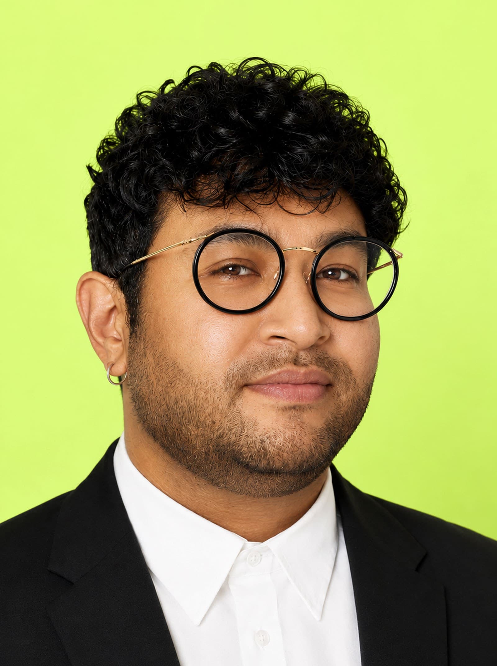 A person with short curly hair, a trimmed beard, and round glasses wears a black suit and white shirt, standing against a bright green background.