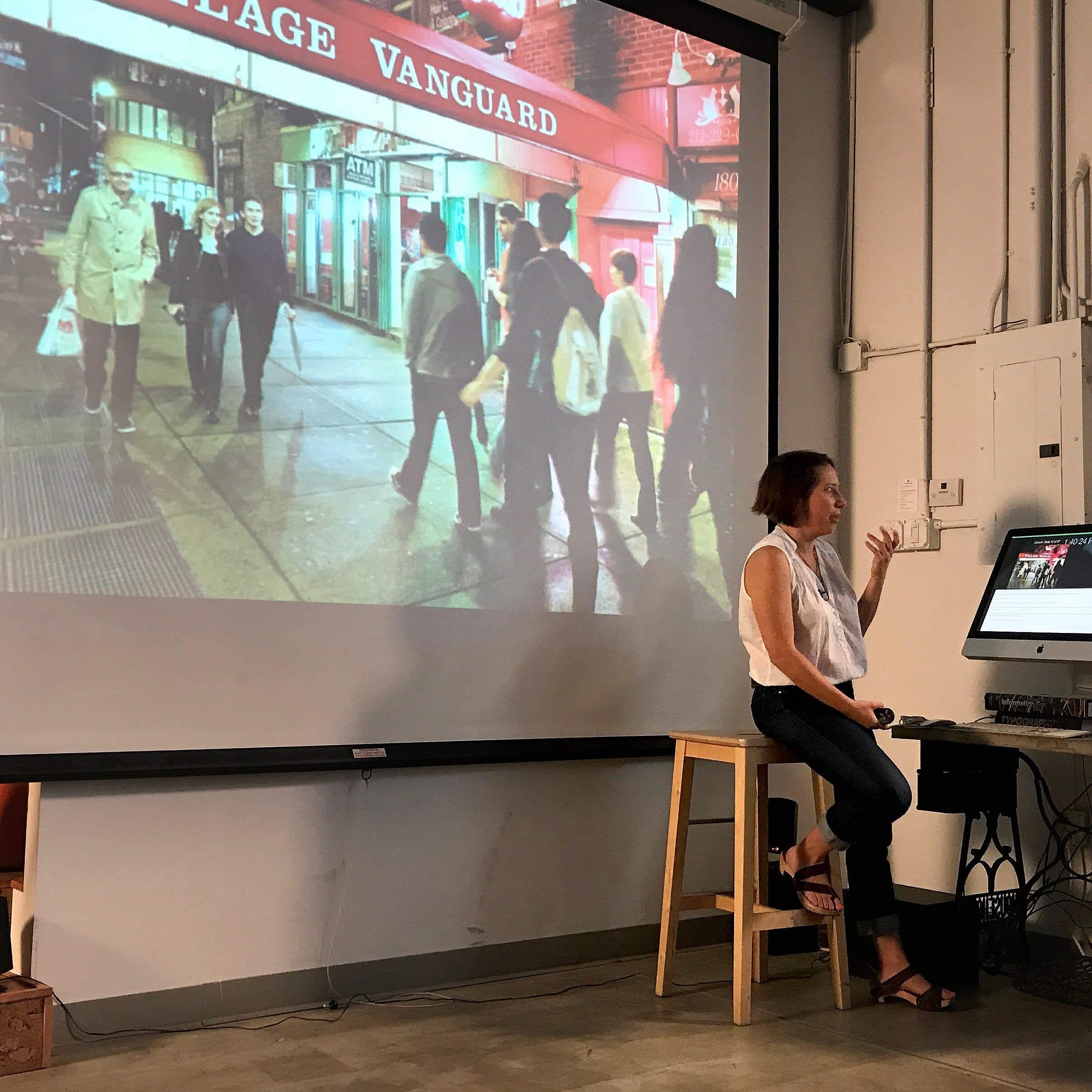 A person is seated on a stool and giving a presentation, gesturing with one hand towards a large photo projected on a screen behind them. The photo depicts a night scene outside the Village Vanguard jazz club, with several people walking on the sidewalk.