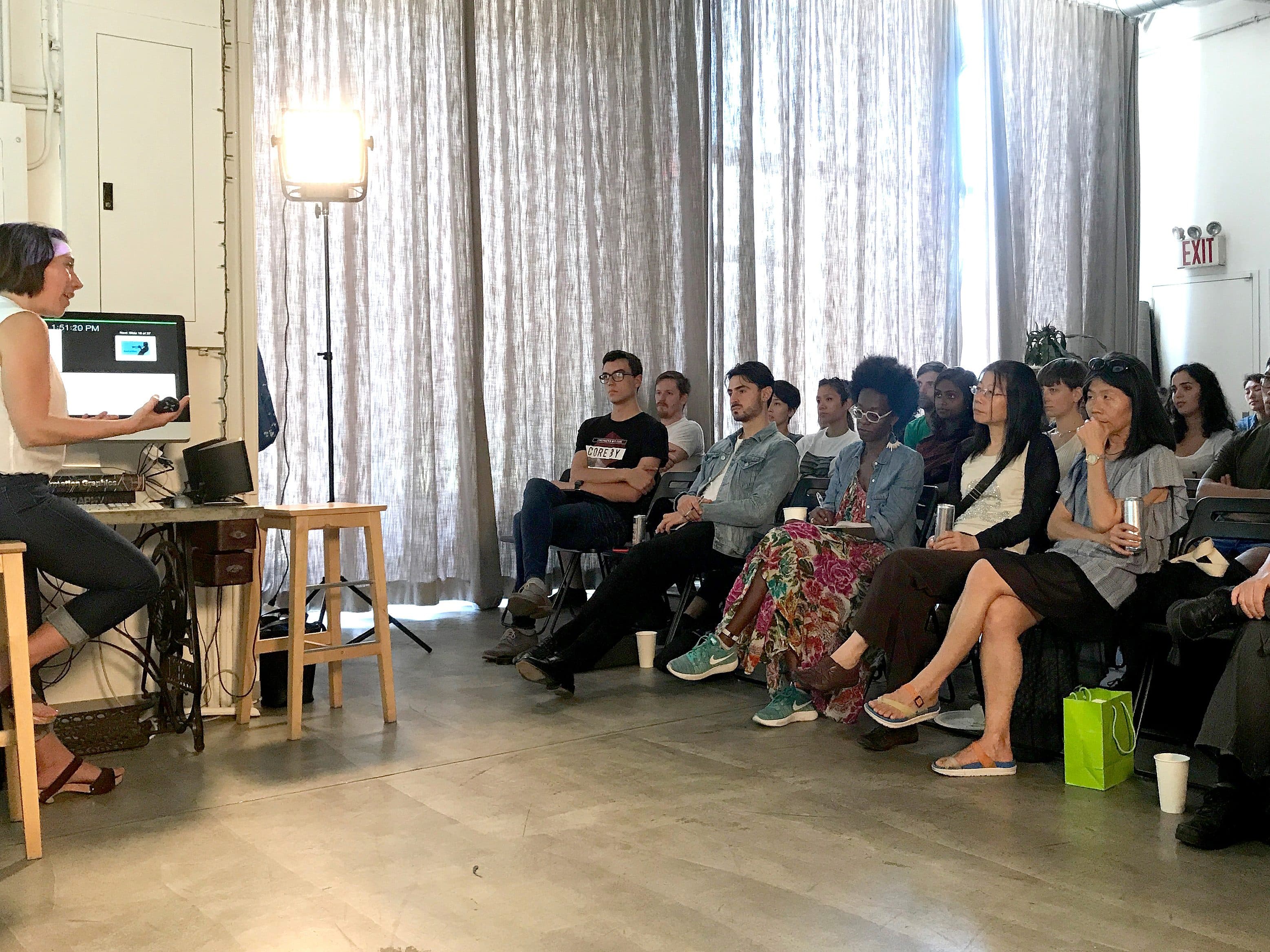 A woman sitting on a stool is giving a presentation to a group of people seated in chairs. There is a laptop and monitor on a small table next to her. The audience is attentively watching, and the room has large windows with curtains allowing natural light.