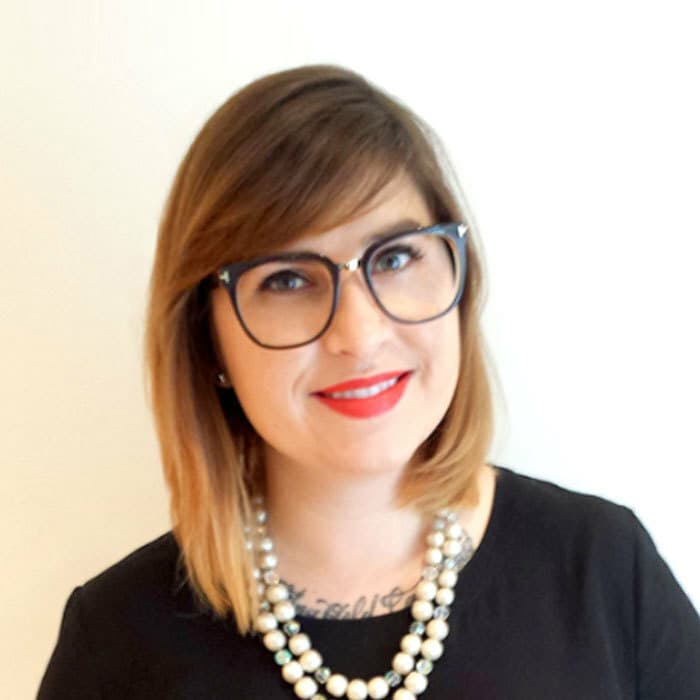 A woman with shoulder-length brown hair, wearing black-framed glasses, a black top, and a pearl necklace, smiles while standing against a plain white background. She has a tattoo partially visible above her neckline.