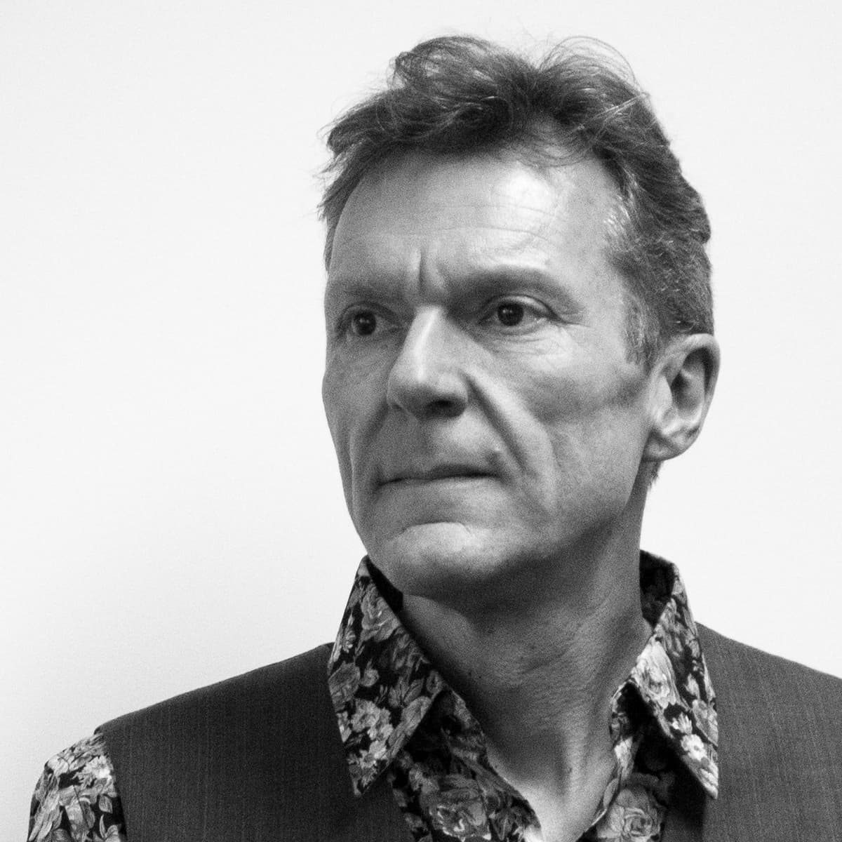 Black and white photo of a middle-aged man with short hair looking slightly to the left. He is wearing a floral patterned shirt under a vest. The background is plain and white.