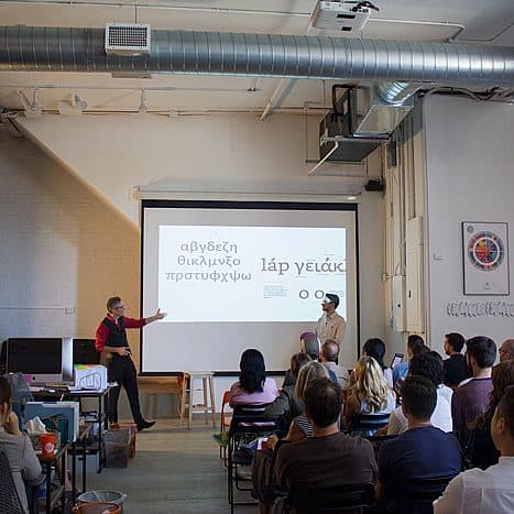 A group of people are seated in a room attentively listening to a presentation. Two presenters stand at the front, gesturing towards a large screen displaying text. The space has an industrial look with exposed ducts and ample natural light from large windows.