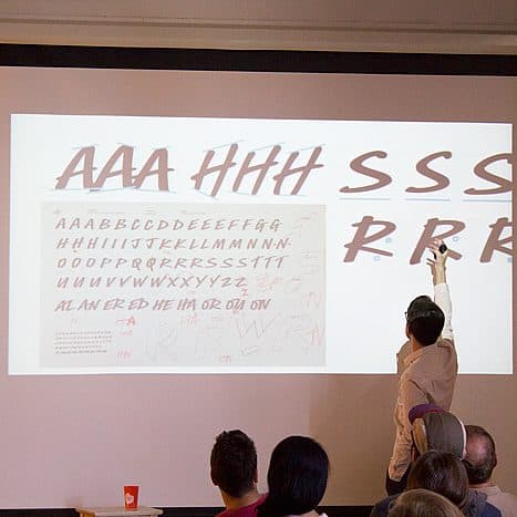 A man is pointing at a projected screen displaying various handwritten letters including "A," "H," "S," and "R" in red ink. Several people are seated facing the screen, and a man in a red shirt stands nearby.