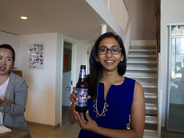 A woman in a blue dress and glasses smiles while holding a beer bottle. Another person beside her, wearing a gray blazer, seems to be in conversation. The background shows a stairway and a partially open door in a modern, well-lit interior space.