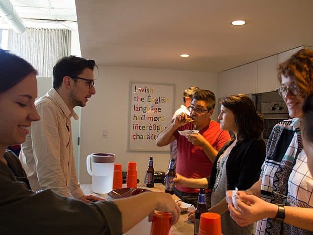 A group of six people stand around a kitchen counter, engaged in conversation and enjoying food and drinks. Red plastic cups and beer bottles are on the counter. In the background, a sign with colorful text reads, "I wish the English language had more etymological characters.