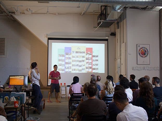 A presenter in a pink shirt stands in front of a projection screen showing a diagram, addressing an audience seated in a room with white walls and industrial decor. Another person in a white shirt stands to the side, listening.