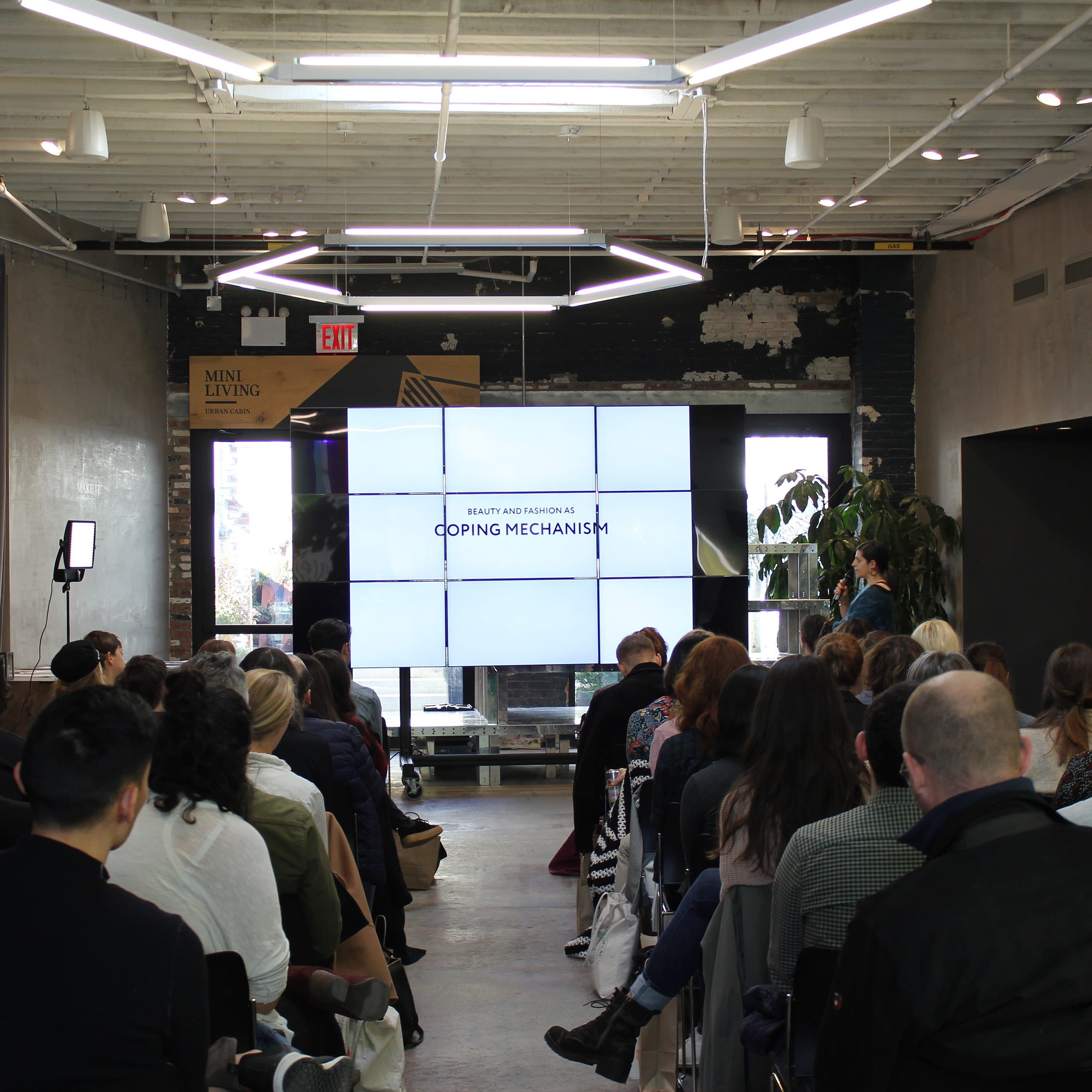 A group of people seated in rows inside a modern industrial-style room with high ceilings and exposed beams, facing a large screen displaying the title "COPING MECHANISMS". The audience appears engaged, listening to the speaker standing next to the screen.