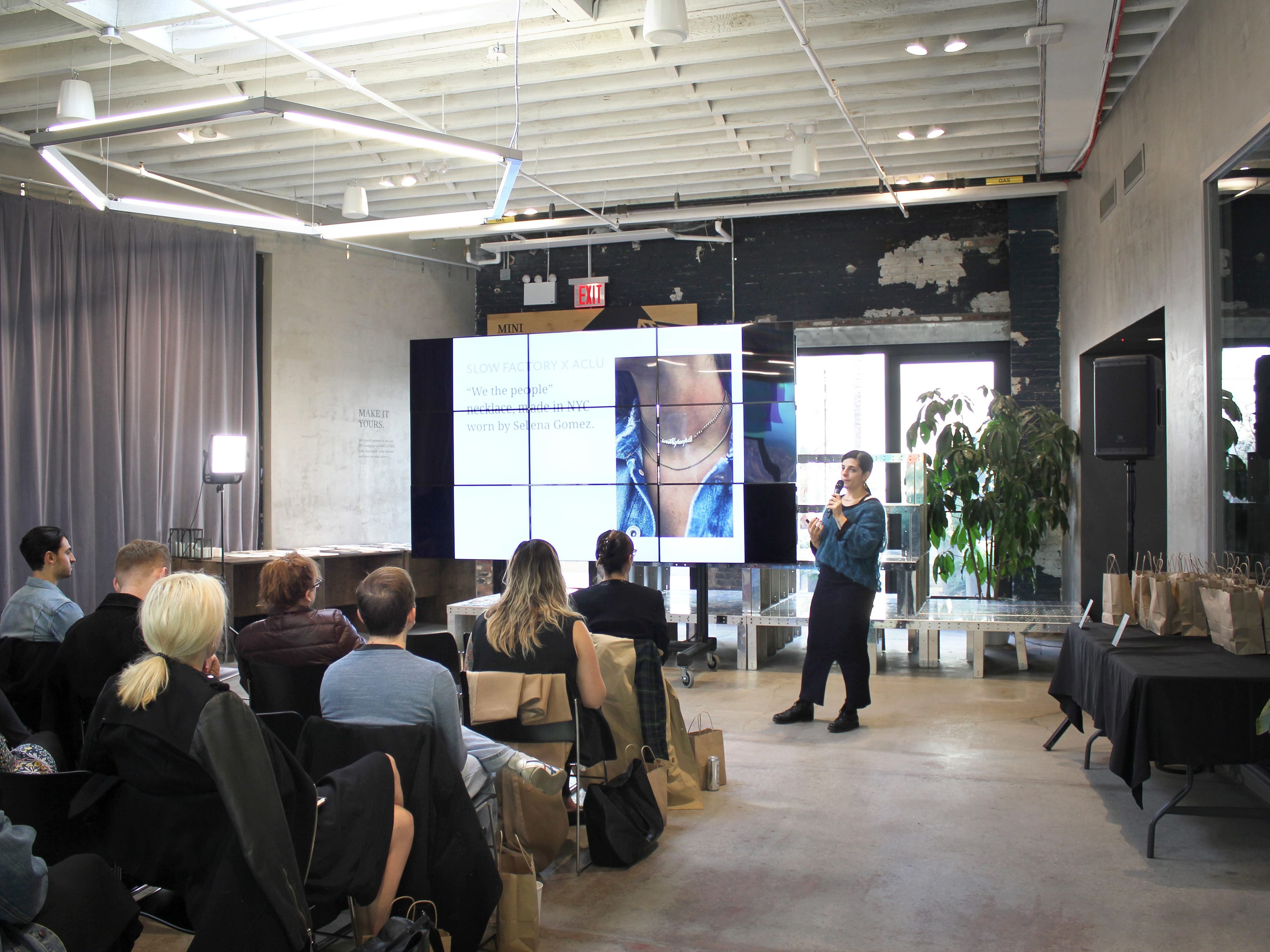 A speaker presents to an audience in a modern, industrial-style room. The audience is seated and facing a large screen displaying a jewelry ad. Tables with gift bags are placed on the right side of the room. The room has large windows, exposed brick, and high ceilings.
