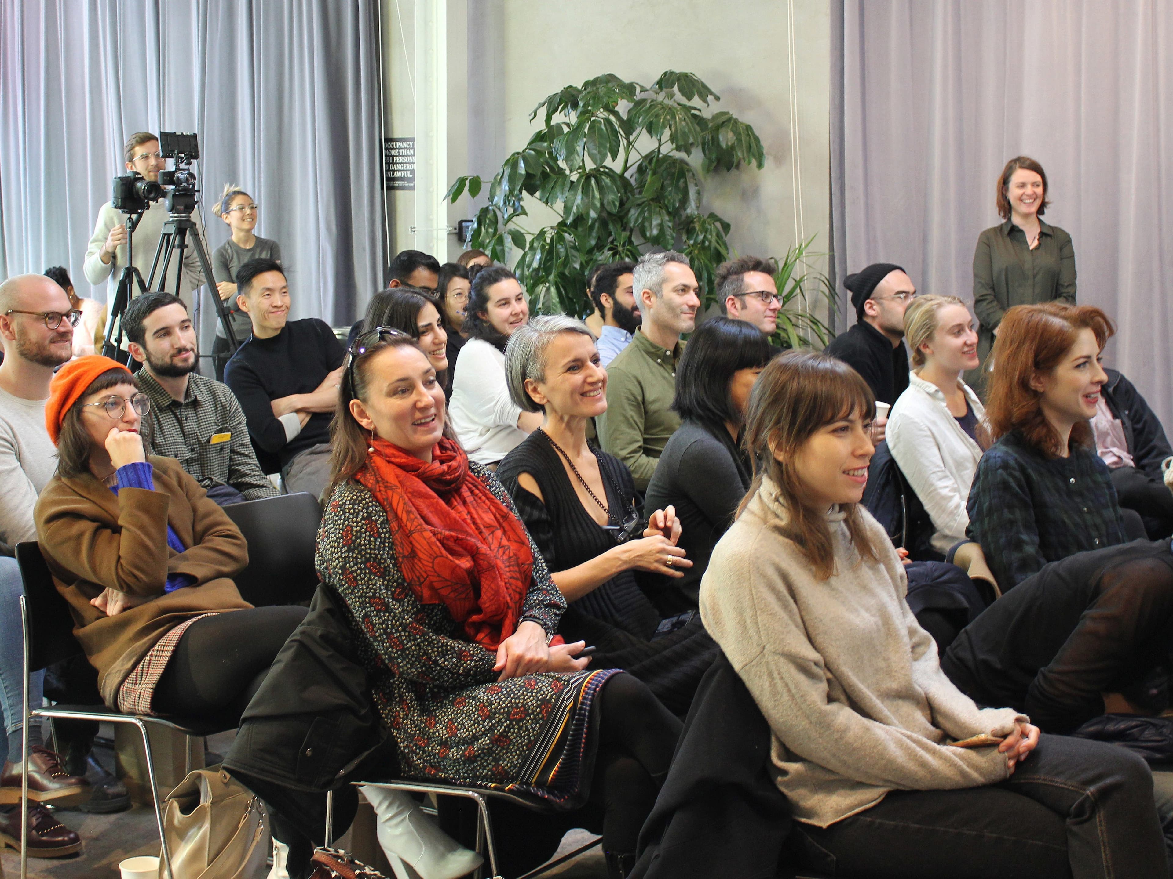 A diverse group of people sit attentively in rows of chairs, watching something off-camera. Some are smiling and laughing. There are two people in the back operating a camera. The setting appears casual and intimate, with natural light and indoor plants.