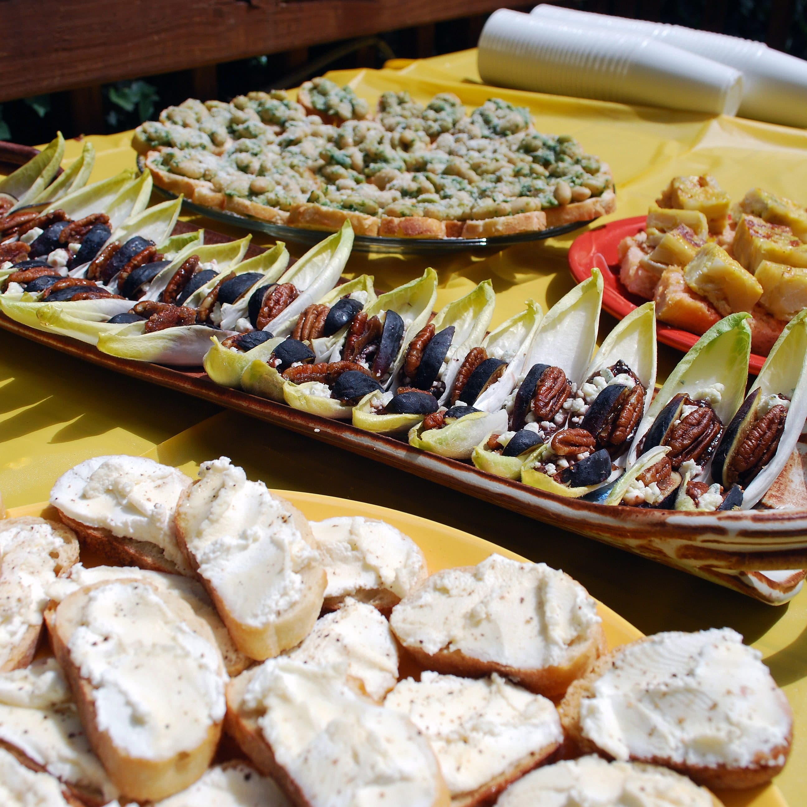 A table is set with an assortment of appetizers. There are slices of bread with a creamy spread, endive leaves topped with nuts and dried fruits, and other various finger foods and snacks. The tablecloth is yellow, and there are several stacked plastic cups in the background.