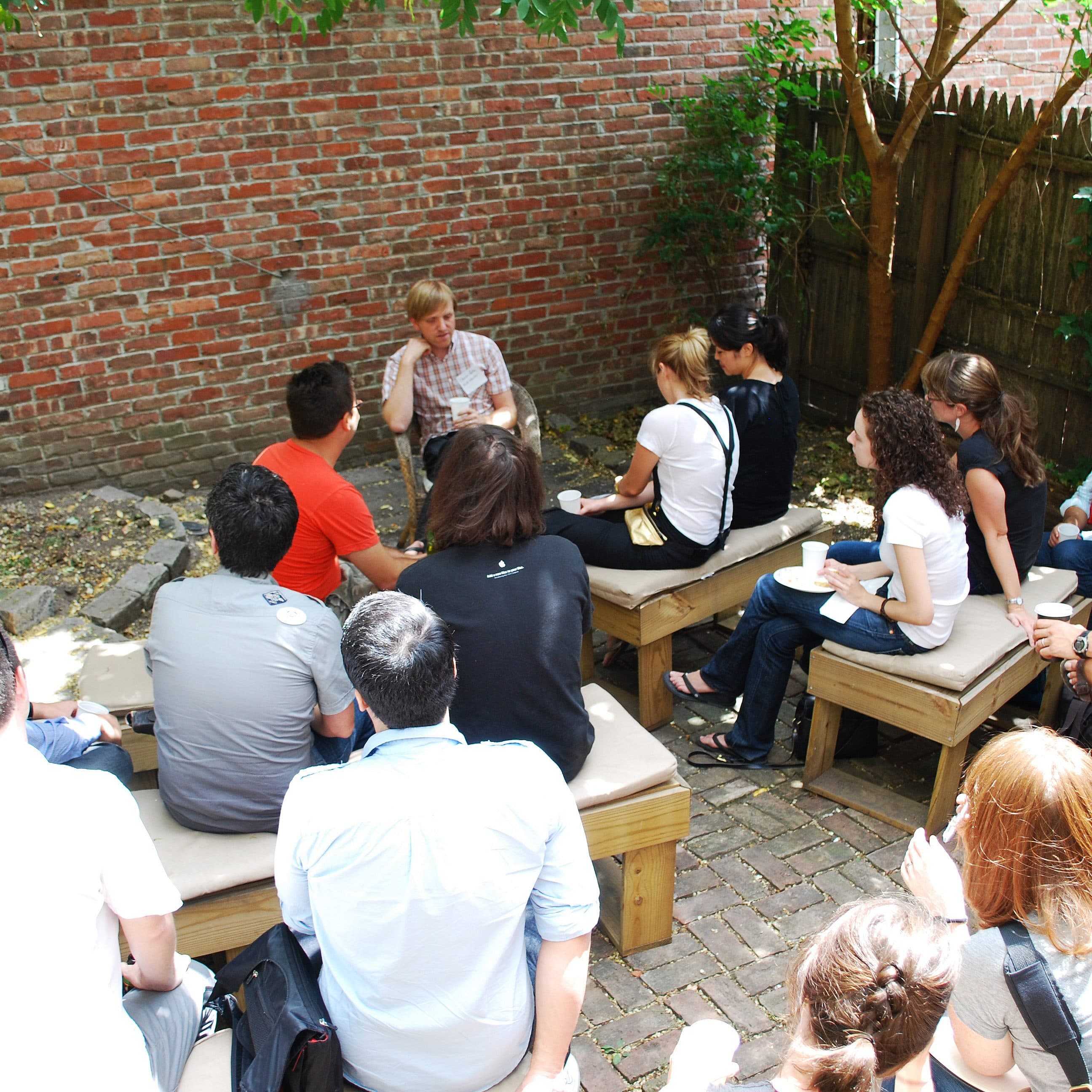 A group of people is seated outside in a courtyard on benches and stools, attentively listening to a person speaking near a brick wall. The setting is casual, with trees providing shade and some greenery around. The speaker is seated, engaged in conversation.