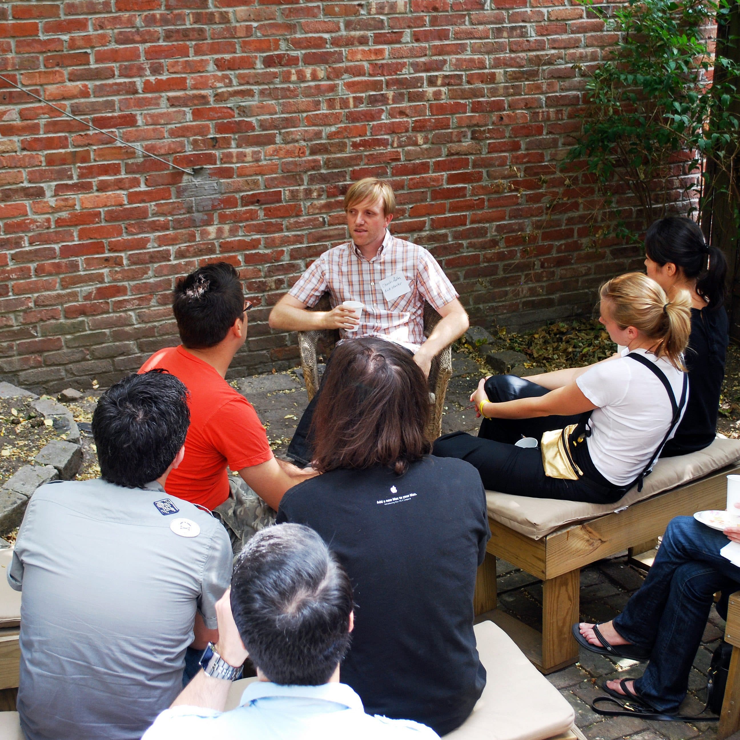 A group of people sit and listen attentively to a person speaking in an outdoor setting with a brick wall and greenery. They are seated on wooden benches arranged in a semicircle, and the speaker appears to be explaining something or leading a discussion.