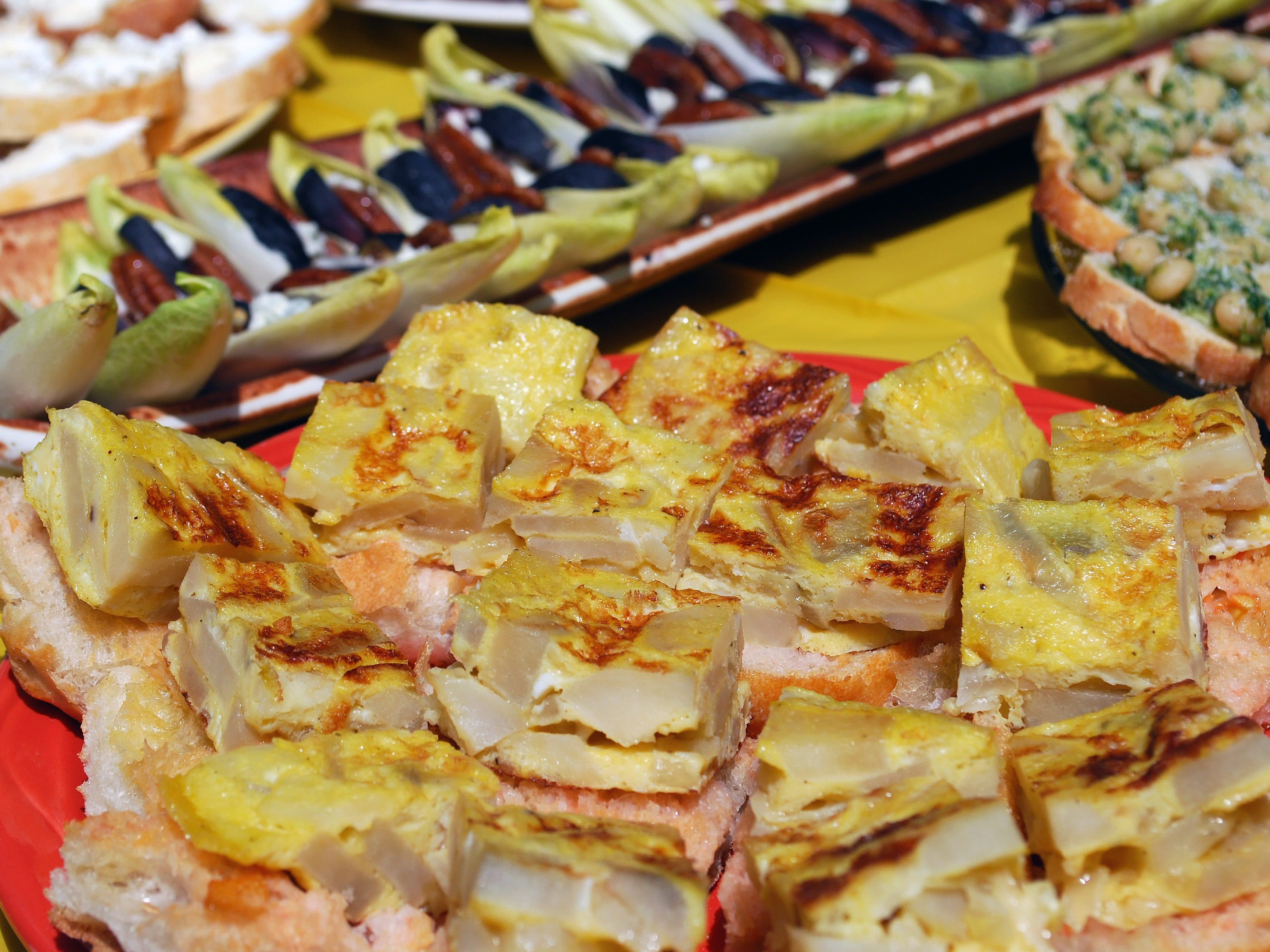 A plate with several small squares of Spanish tortilla (a potato and egg dish) placed on top of sliced bread. In the background are other assorted appetizers, including endive leaves with toppings and bread with white beans. The tablecloth is bright yellow.