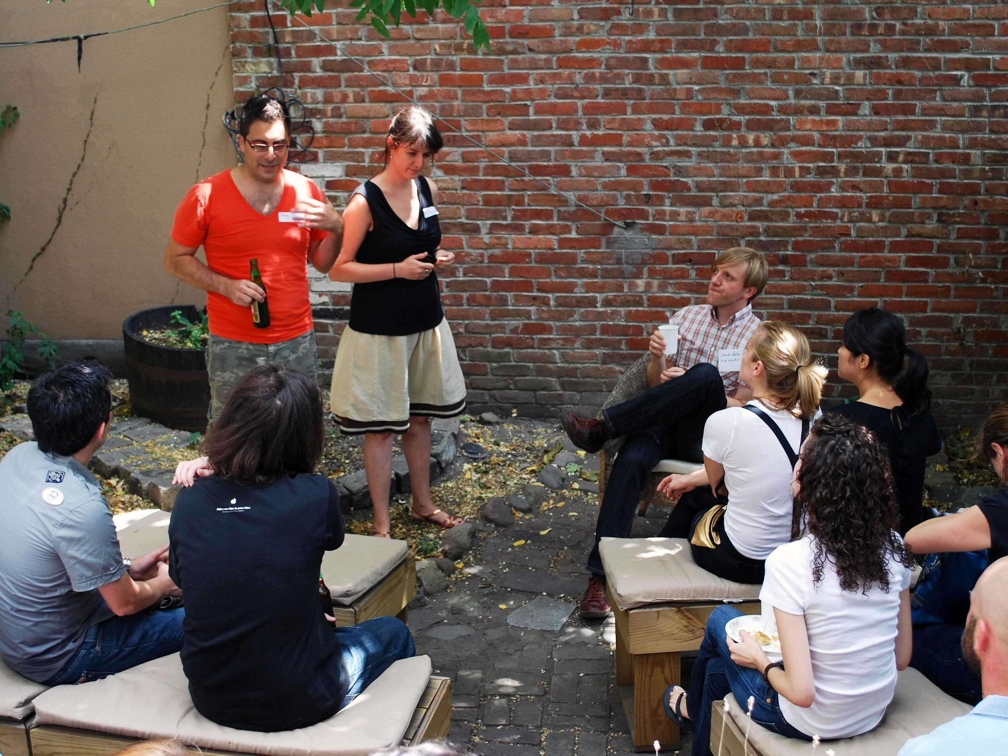 A group of people are gathered in an outdoor setting with a brick wall background. Two individuals are standing, one holding a beer and speaking, while the others are seated, listening attentively. Casual clothing and a relaxed atmosphere suggest an informal event.