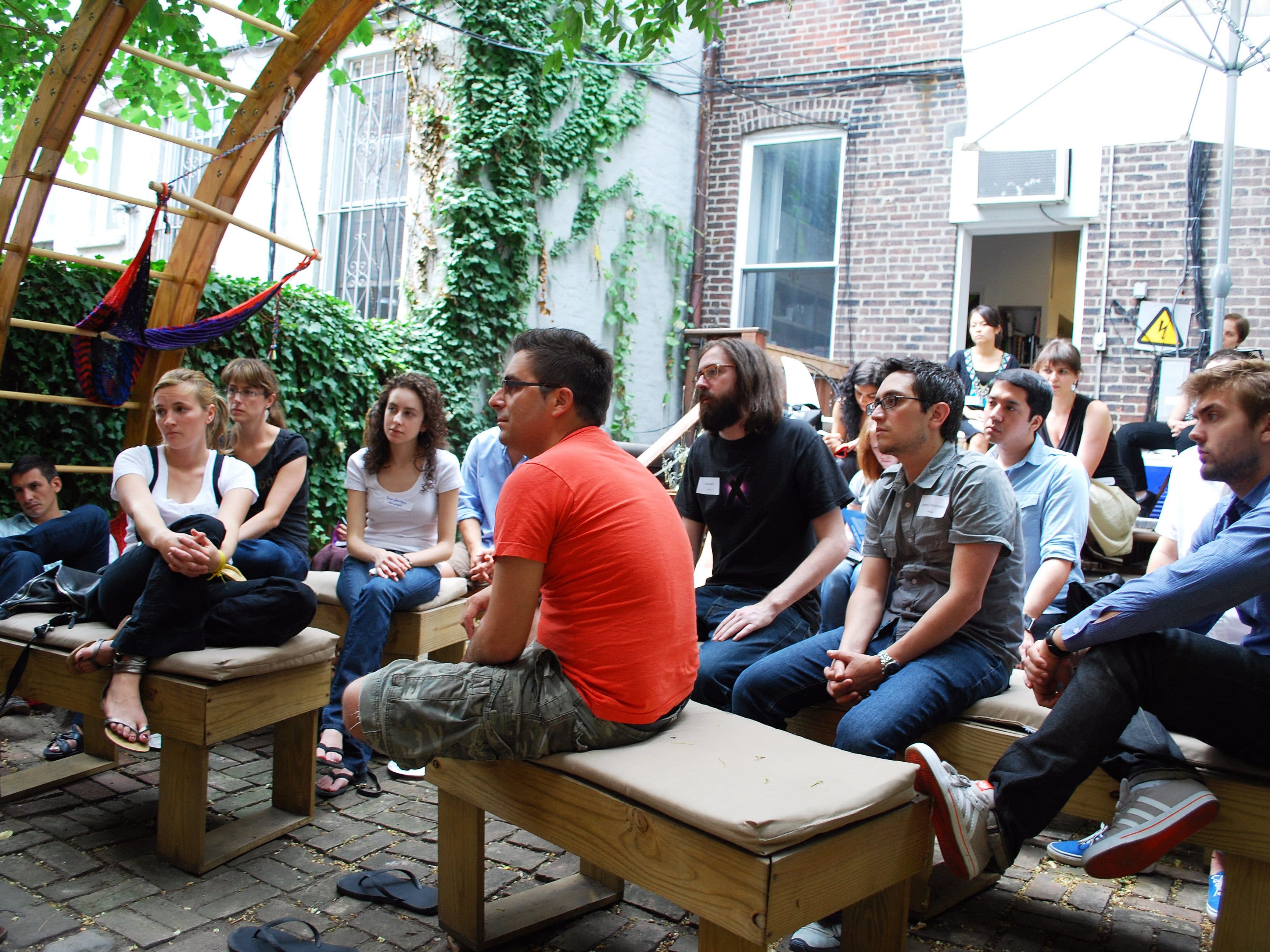 A group of people sit on benches outside in a courtyard surrounded by ivy-covered brick walls. One person in the foreground is wearing a red shirt, while others are dressed casually. They appear to be attentively listening to someone.