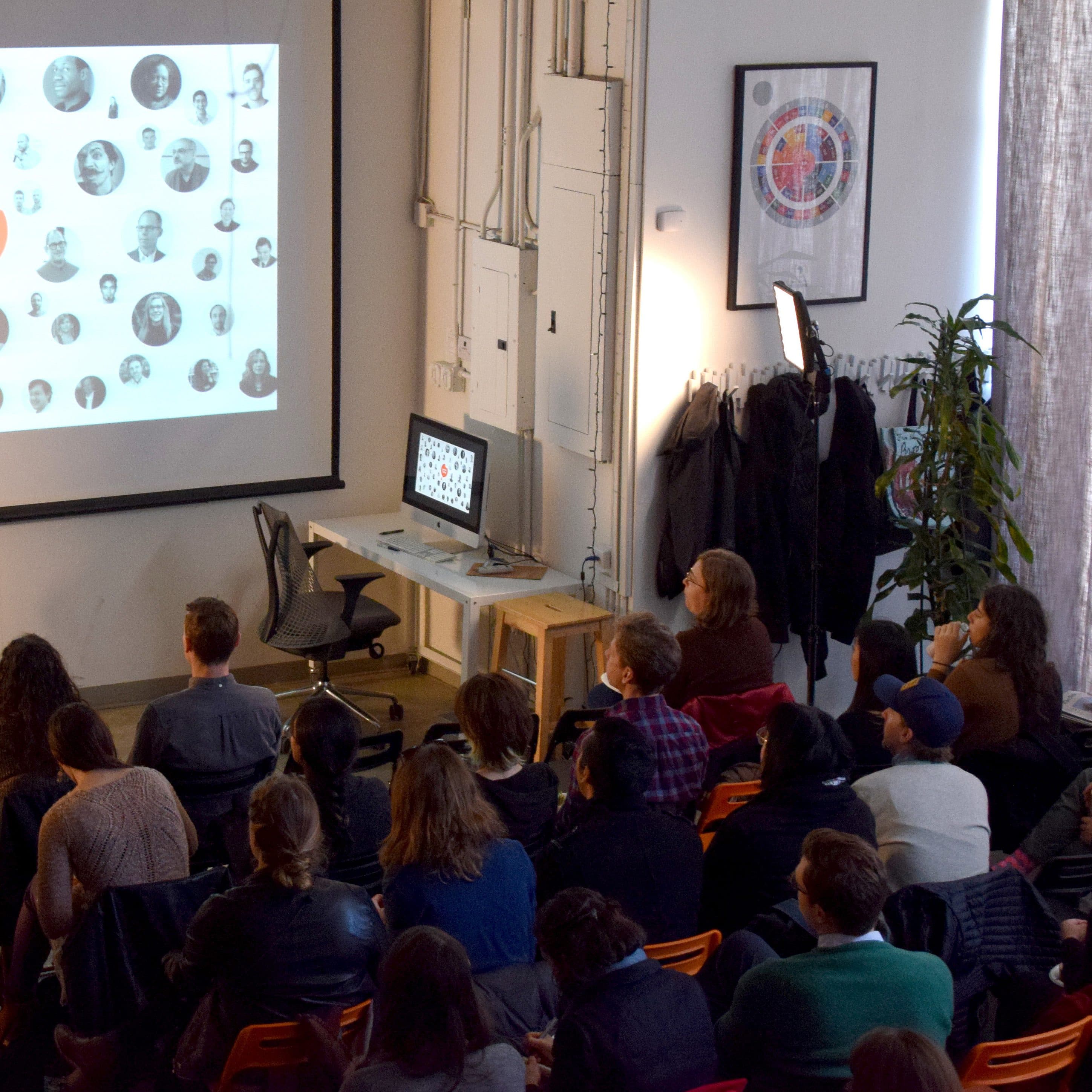 A presenter stands next to a screen displaying various black-and-white headshots and text that reads "Lunch Talks." The audience, seated in rows, attentively watches. The setting is a modern room with a computer on a desk, a potted plant, and coats hanging on a rack.