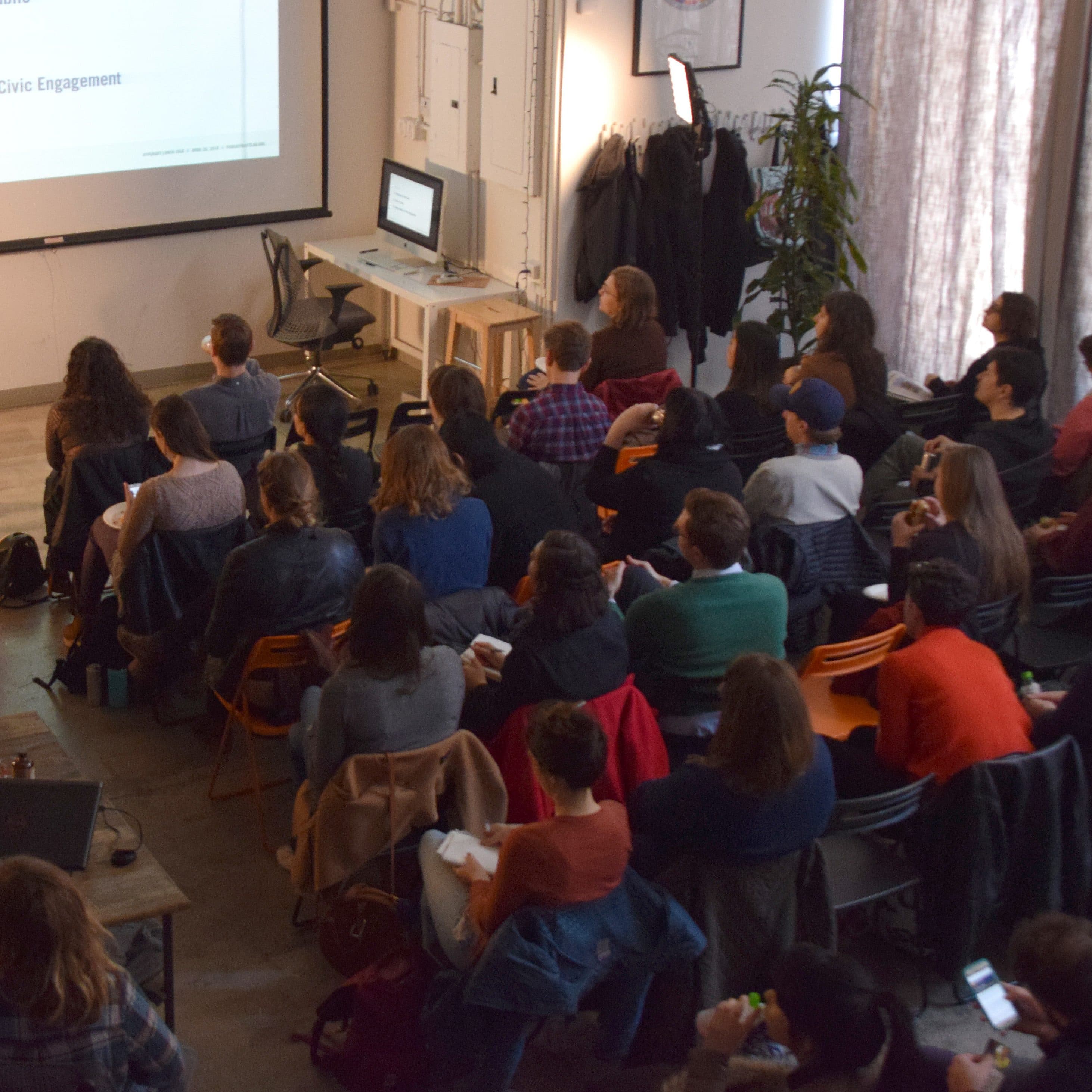 A woman in an orange dress is presenting to an attentive audience in a modern conference room. The audience is seated on chairs and some are holding notebooks. The presentation slide on the screen lists topics related to design and ethics.