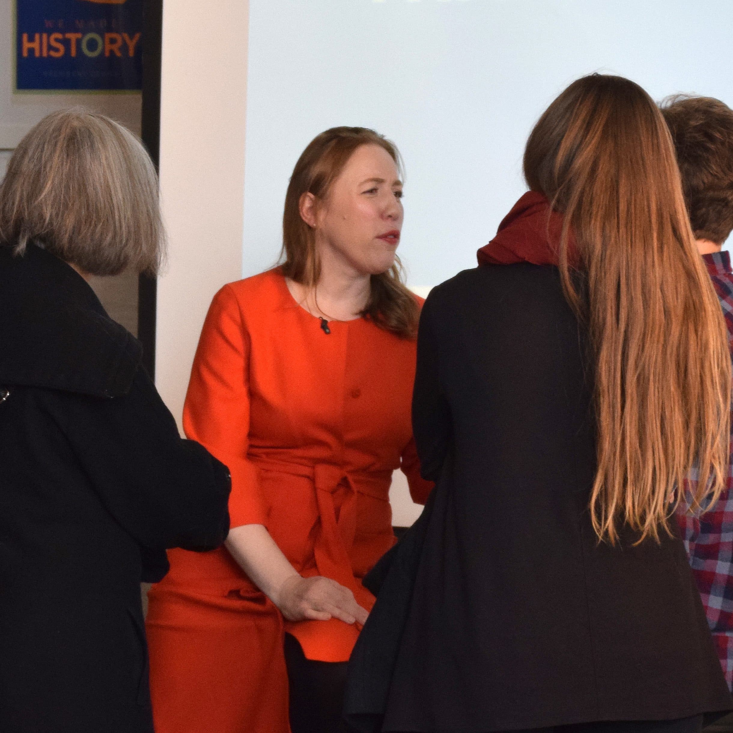 A woman in an orange dress is speaking with three people in a dimly lit room. Two individuals have long hair and one person is wearing a checkered shirt. A projection screen is in the background with posters that have the word "HISTORY" partially visible.