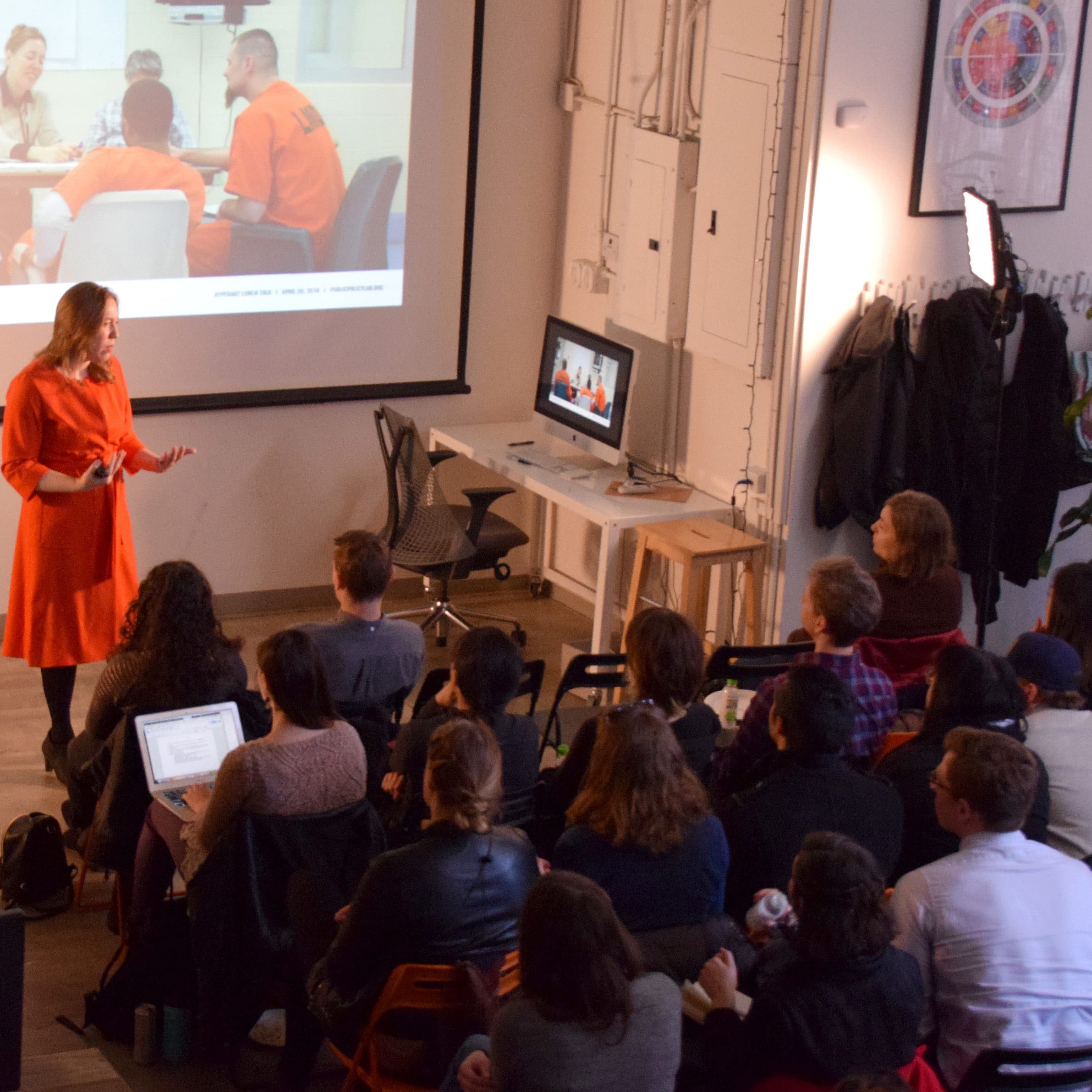 A person stands in front of a room of seated people, presenting a slideshow with an image of individuals in orange uniforms. The presenter is wearing an orange outfit and gesturing towards the screen. The room has a desk, computer, and coats hanging on hooks.