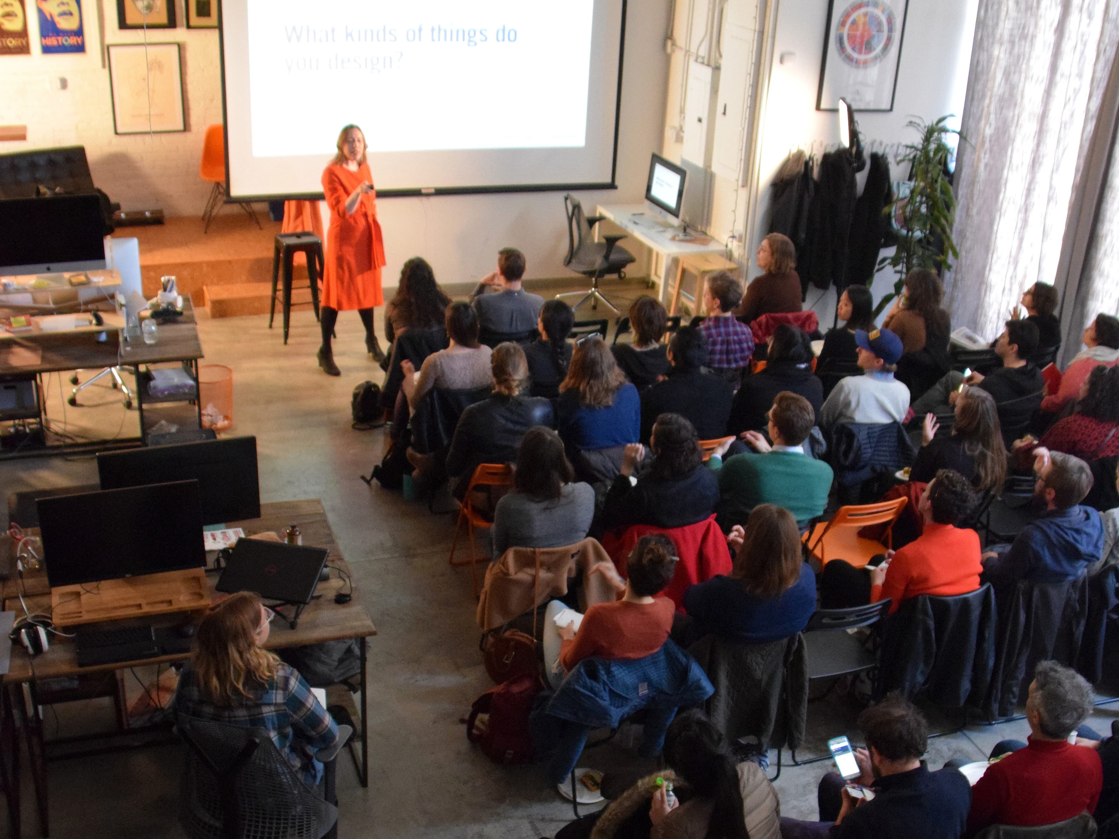 A woman in an orange dress stands at the front of a room, speaking to an audience seated in chairs. The audience is attentive, with some taking notes. A screen behind the speaker displays the text "What kinds of things do you design?" The setting appears to be a modern workspace or seminar room, featuring desks with computer monitors, a bookshelf, and framed artwork on the walls.