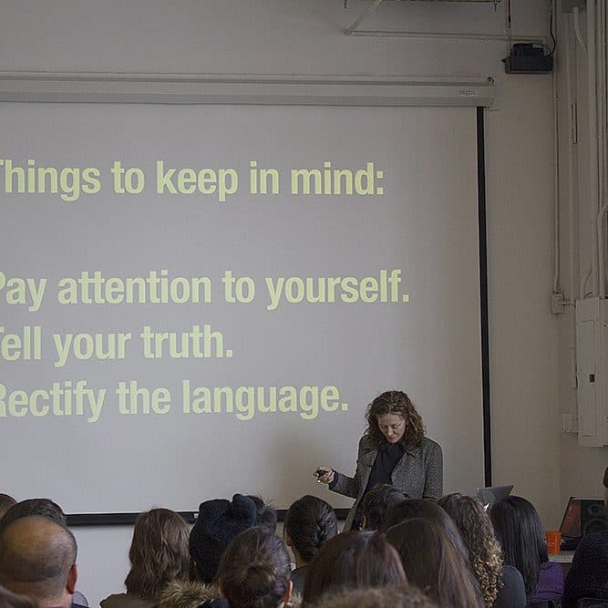 A speaker stands in front of a screen displaying a presentation to a seated audience. The slide on the screen reads: "Things to keep in mind: Pay attention to yourself. Tell your truth. Rectify the language." The room is well-lit and occupied by attentive listeners.