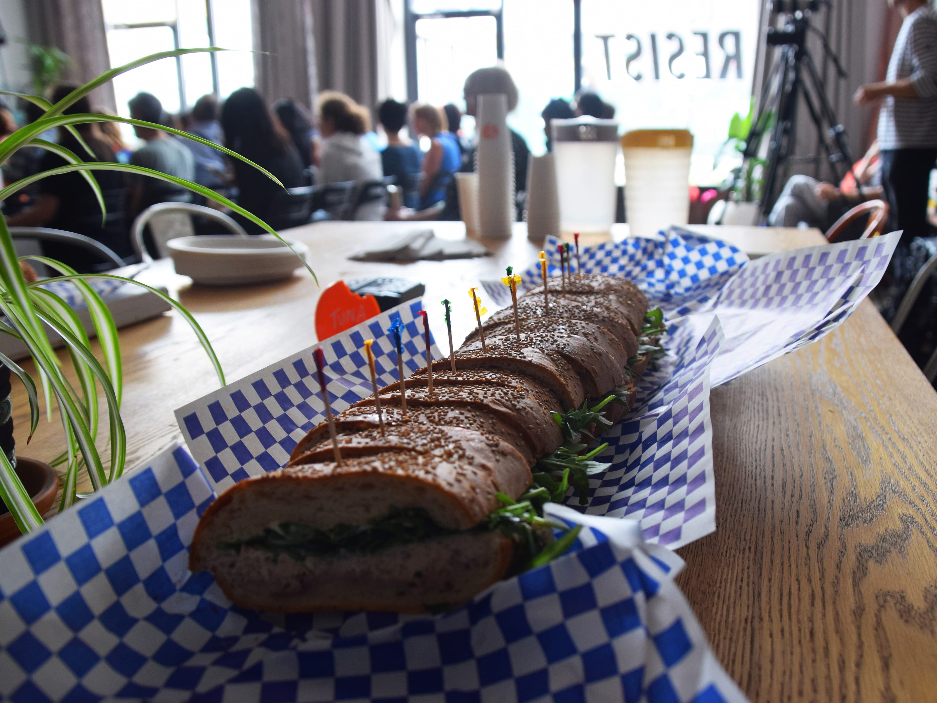A long sandwich sliced into pieces is displayed on a table with blue and white checkered paper. The sandwich has various colored toothpicks stuck into it, and there are some people blurred in the background, suggesting a gathering or meeting. Potted plants and drinks are visible on the table.